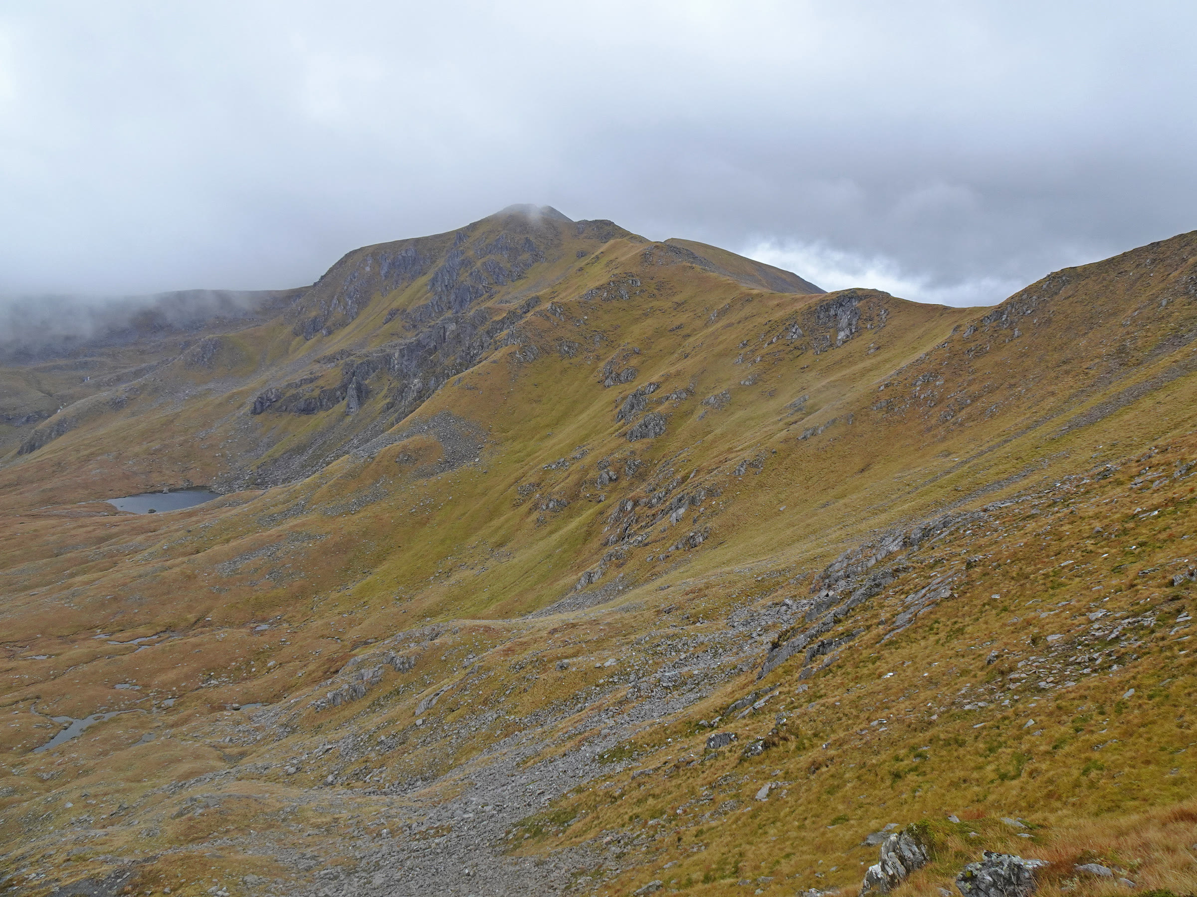 A rounded, crag-streaked mountain with its top in cloud, above a small tarn and open autumn hillside.