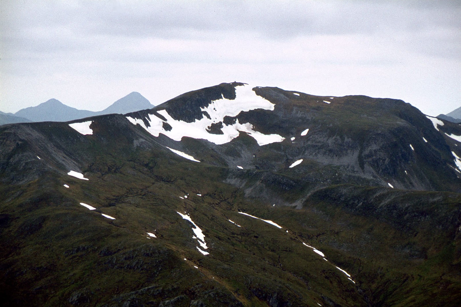 A dark mountain with patches of snow near its summit, seen from a neighbouring peak, with a distant pair of peaks behind.