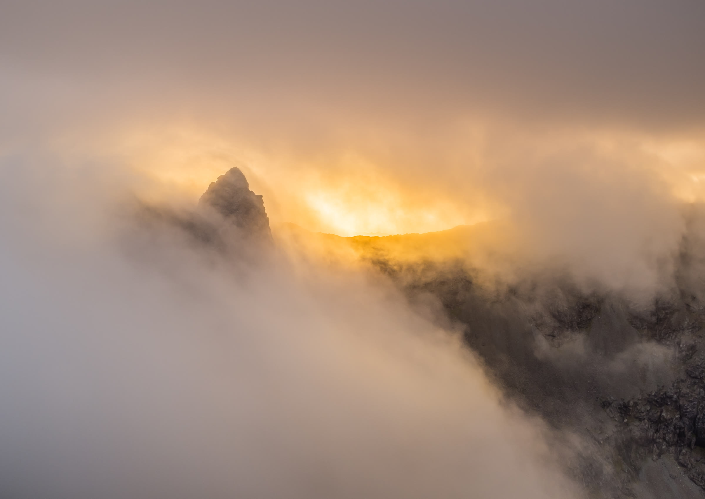 The rock fang of Am Basteir rising out of cloud lit gold by a low sun.