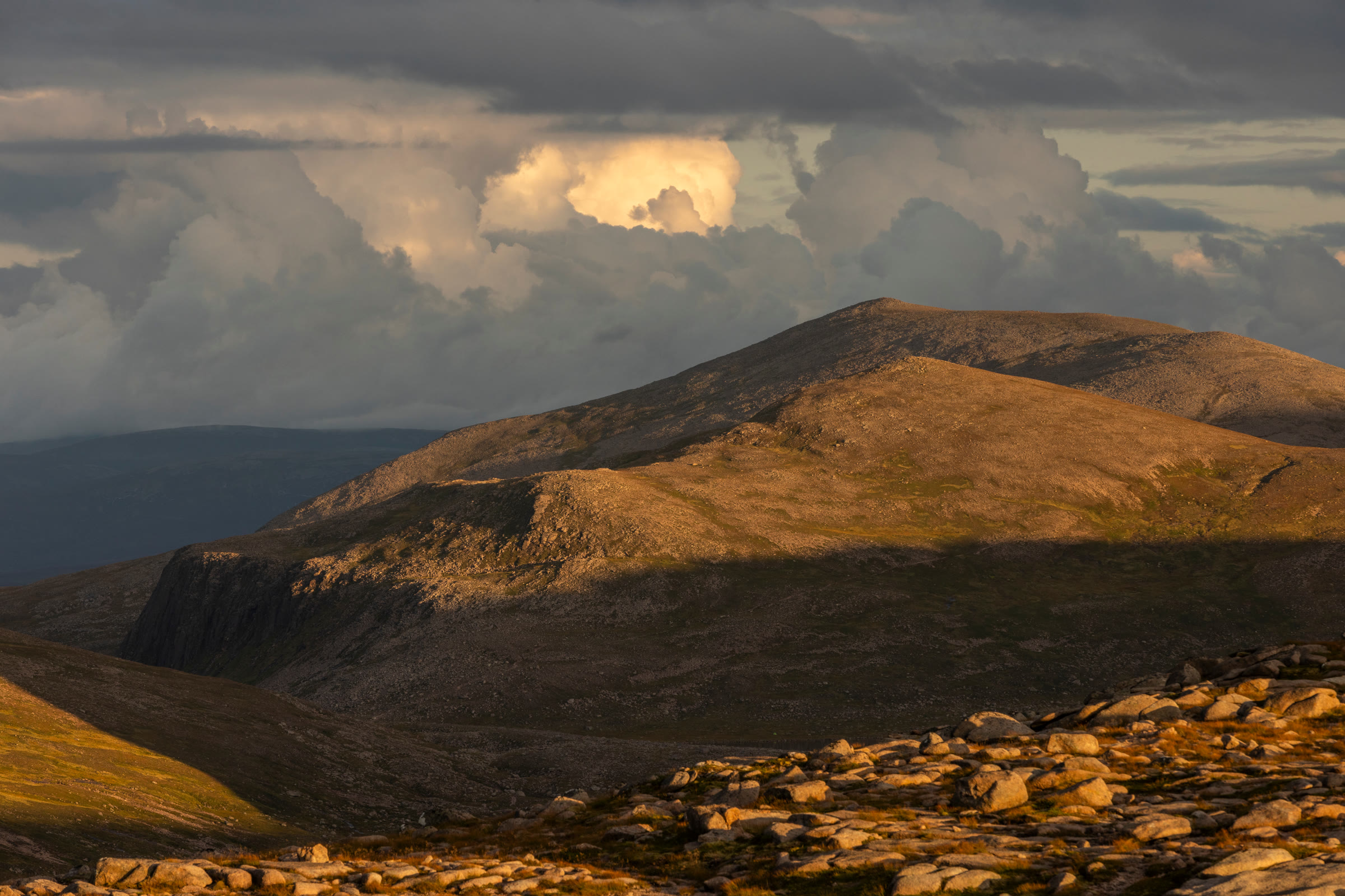 Derry Cairngorm lit copper under towering storm cloud, boulders in the foreground.