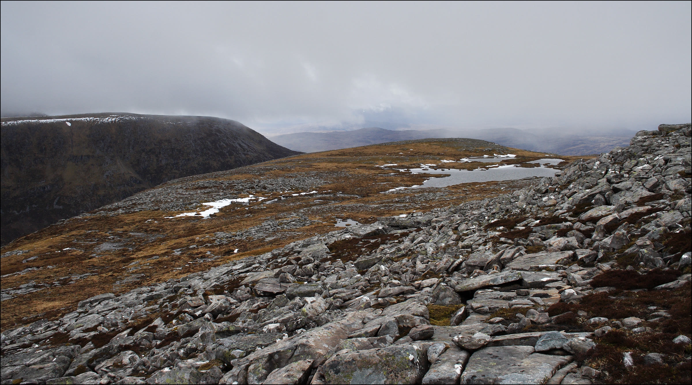 The stony dome of Eididh nan Clach Geala across a bouldery ridge with small lochans and old snow.