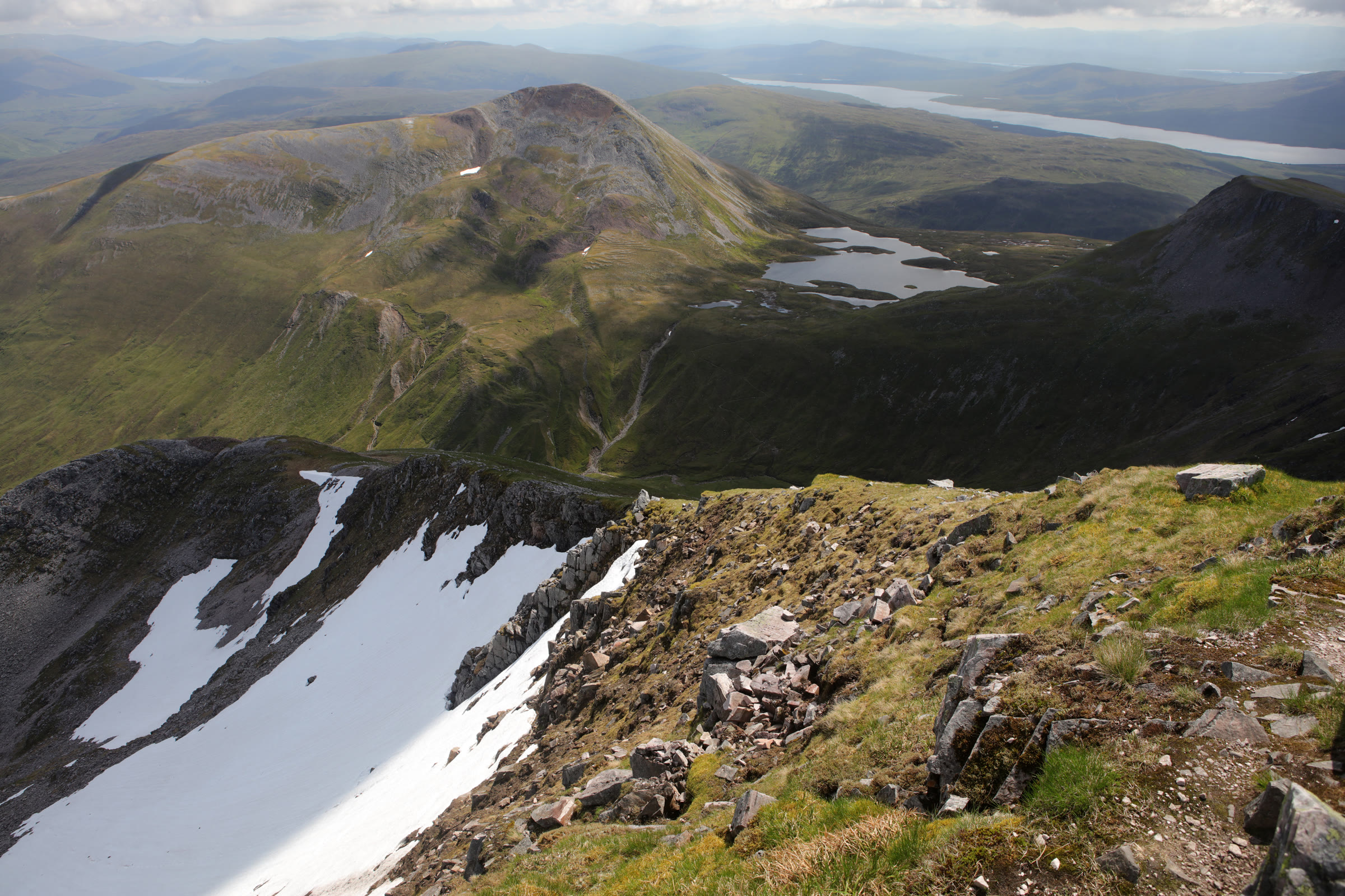 Looking down from the ridge above a snow patch to a scree-sided hill and its lochans, on the way up to Binnein Mòr and Na Gruagaichean.