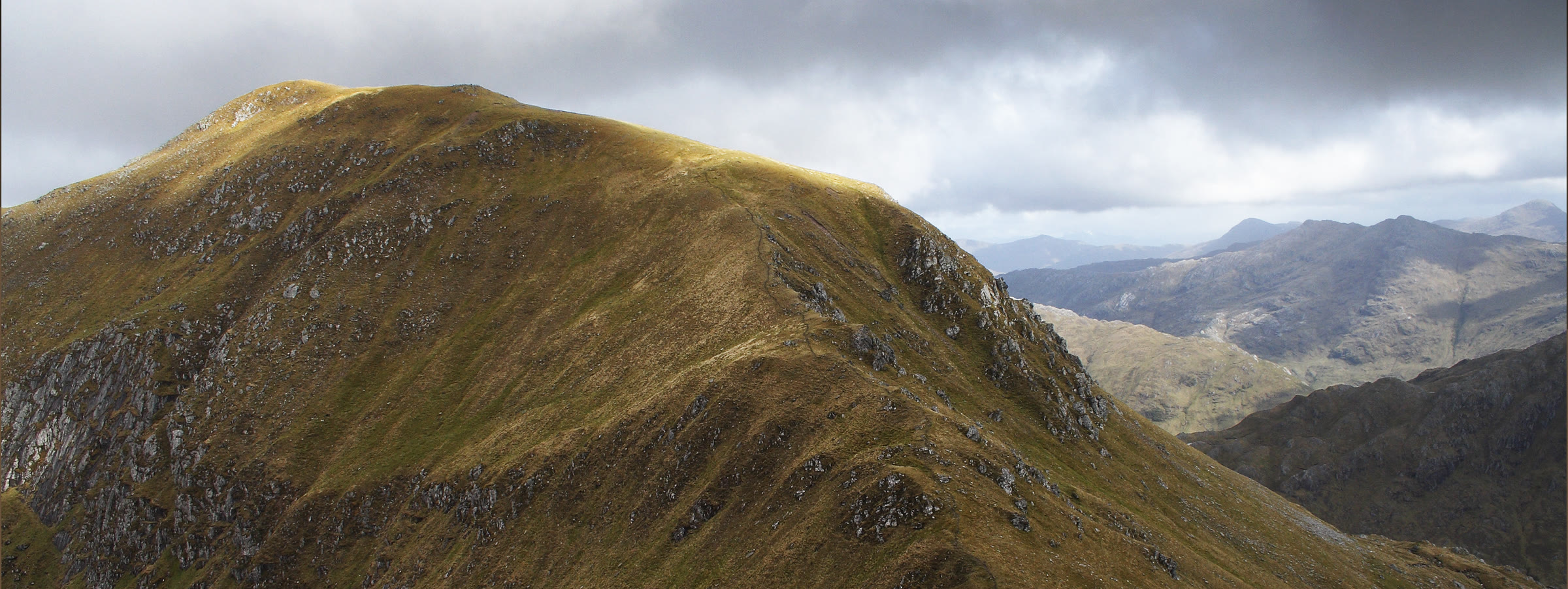 The steep, grassy bulk of Meall Buidhe in sunshine under heavy cloud, the rough hills of Knoydart beyond.