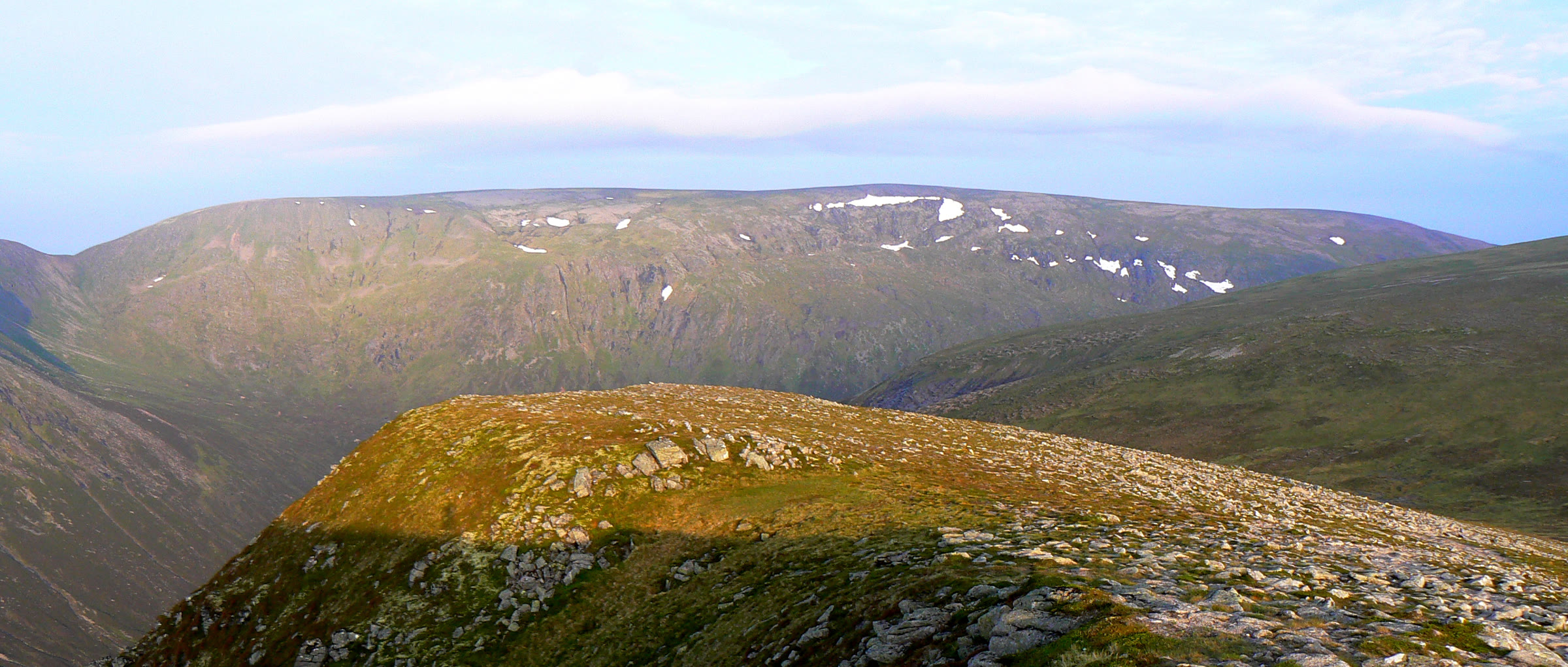The broad plateau of Monadh Mòr with late snow along its rim, beyond a stony ridge in evening light.