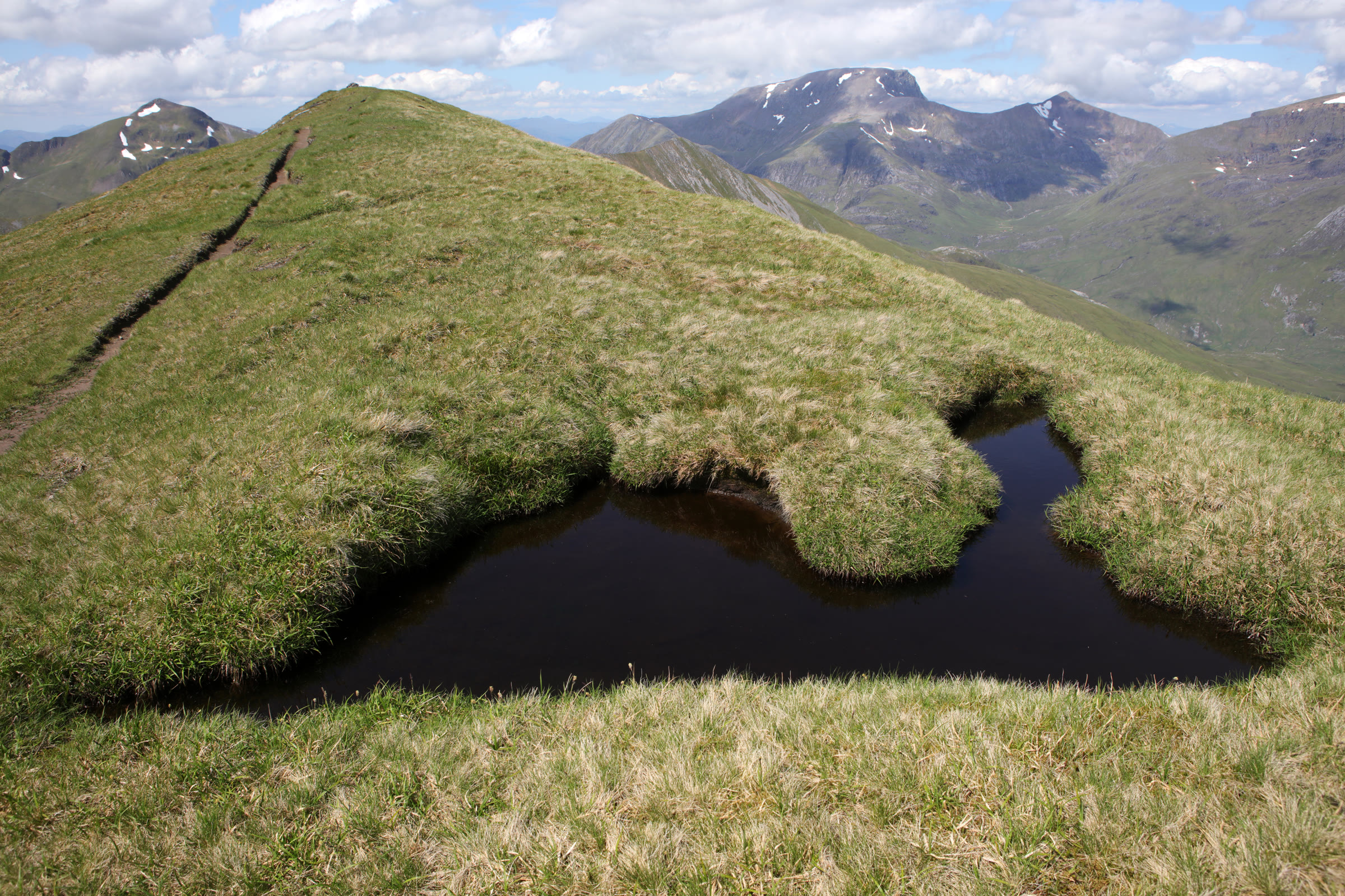 A dark pool on the grassy ridge up to Binnein Mòr and Na Gruagaichean, bigger hills across the glen.