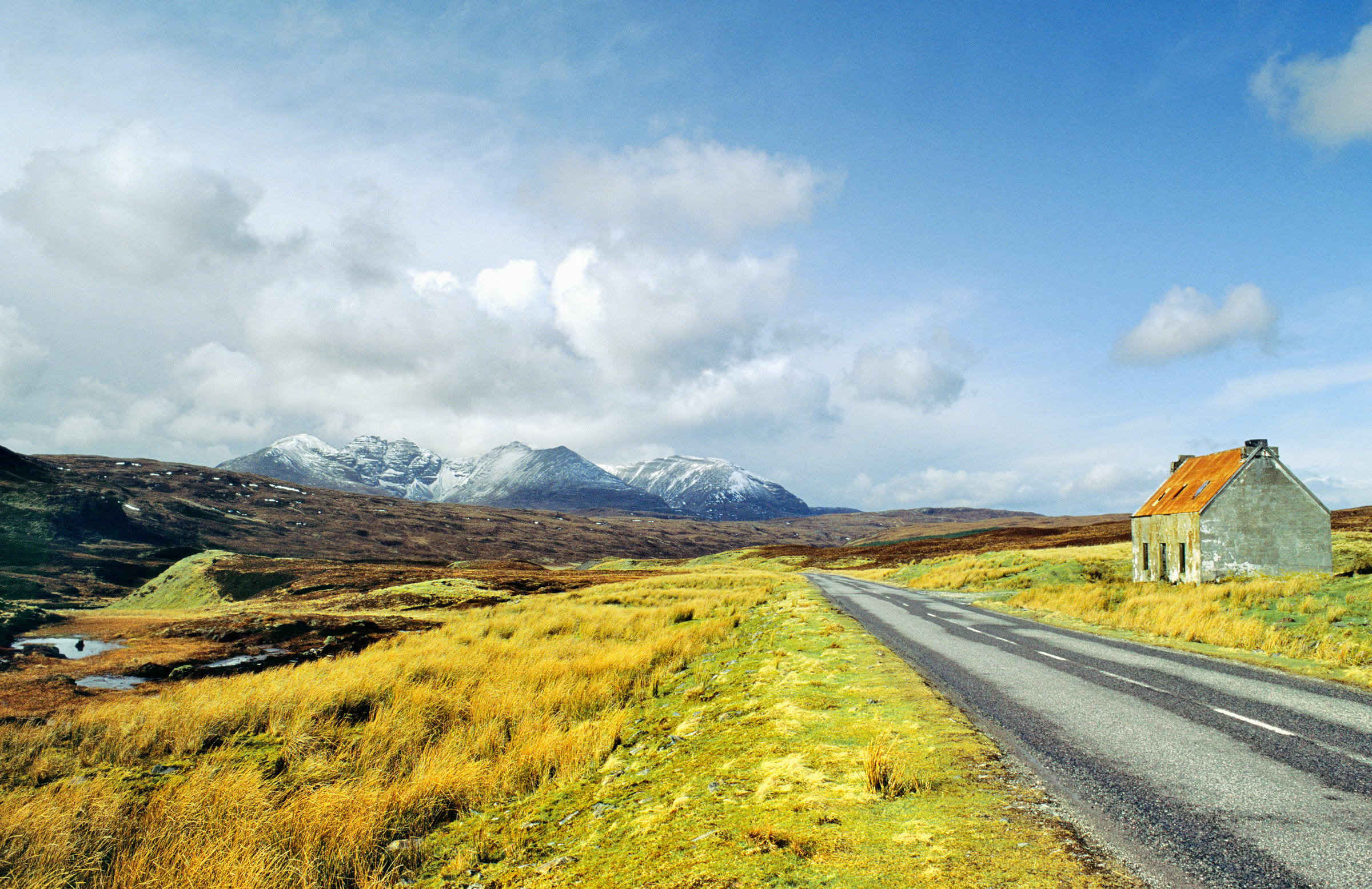A moorland road past a rust-roofed cottage, An Teallach under snow ahead.