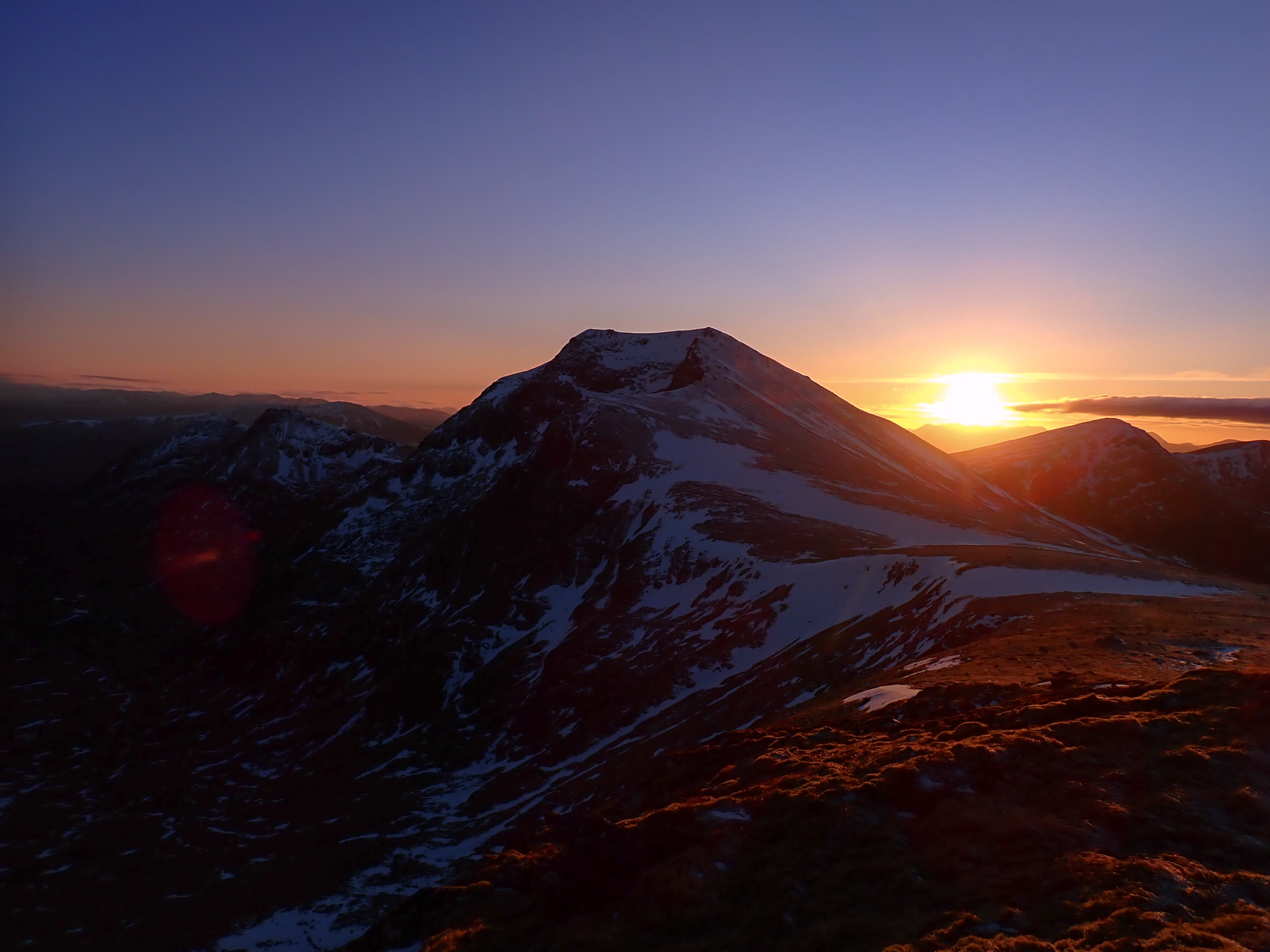Sgùrr nan Clach Geala at sunset, snow streaking its slopes and the sun low beside the summit.