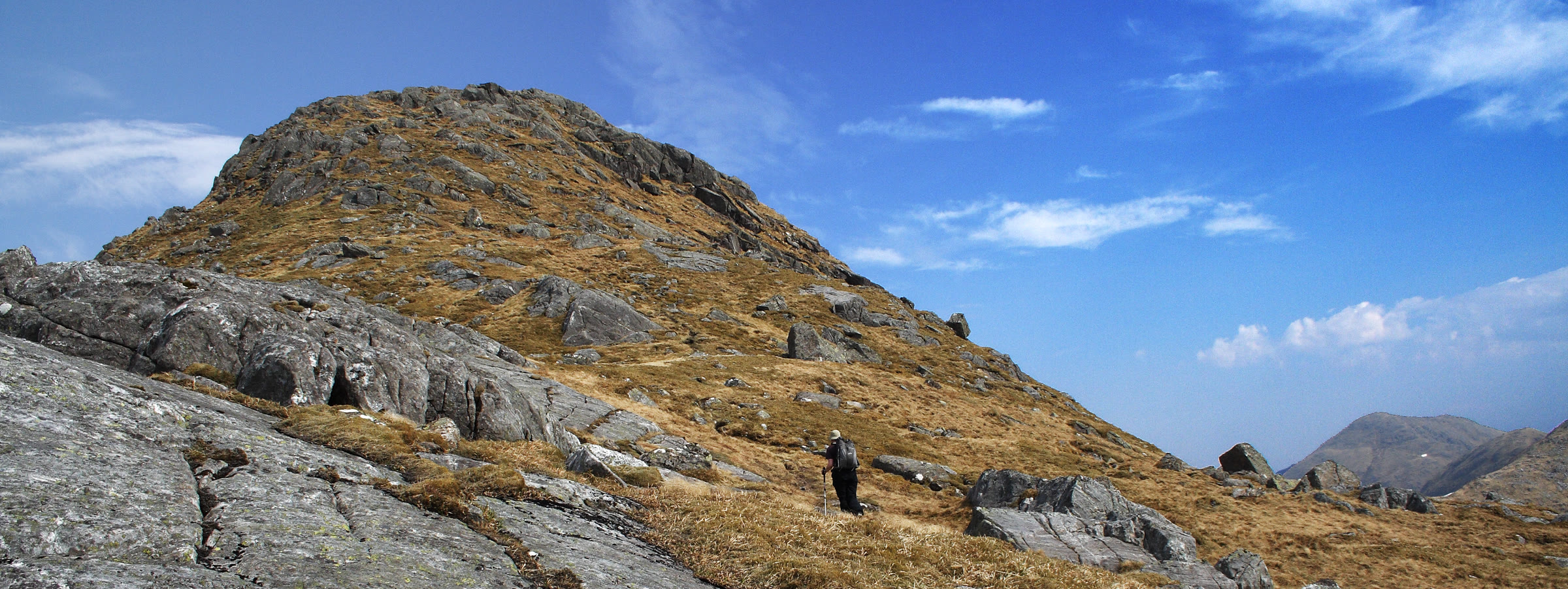 The rocky, grassy top of Sgùrr nan Coireachan under a blue sky, bare slabs in front.