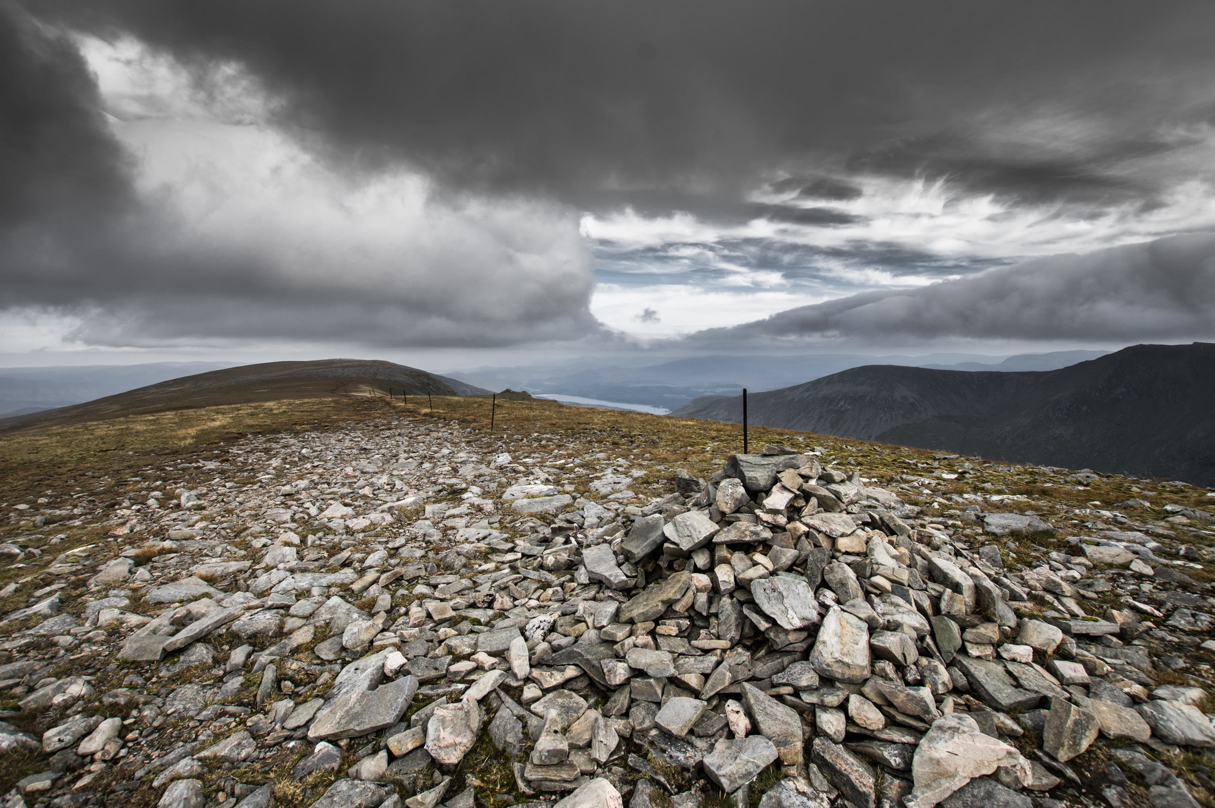 The summit cairn of Stob Poite Coire Àrdair on a stony plateau under heavy cloud, old fence posts running away along the ridge.