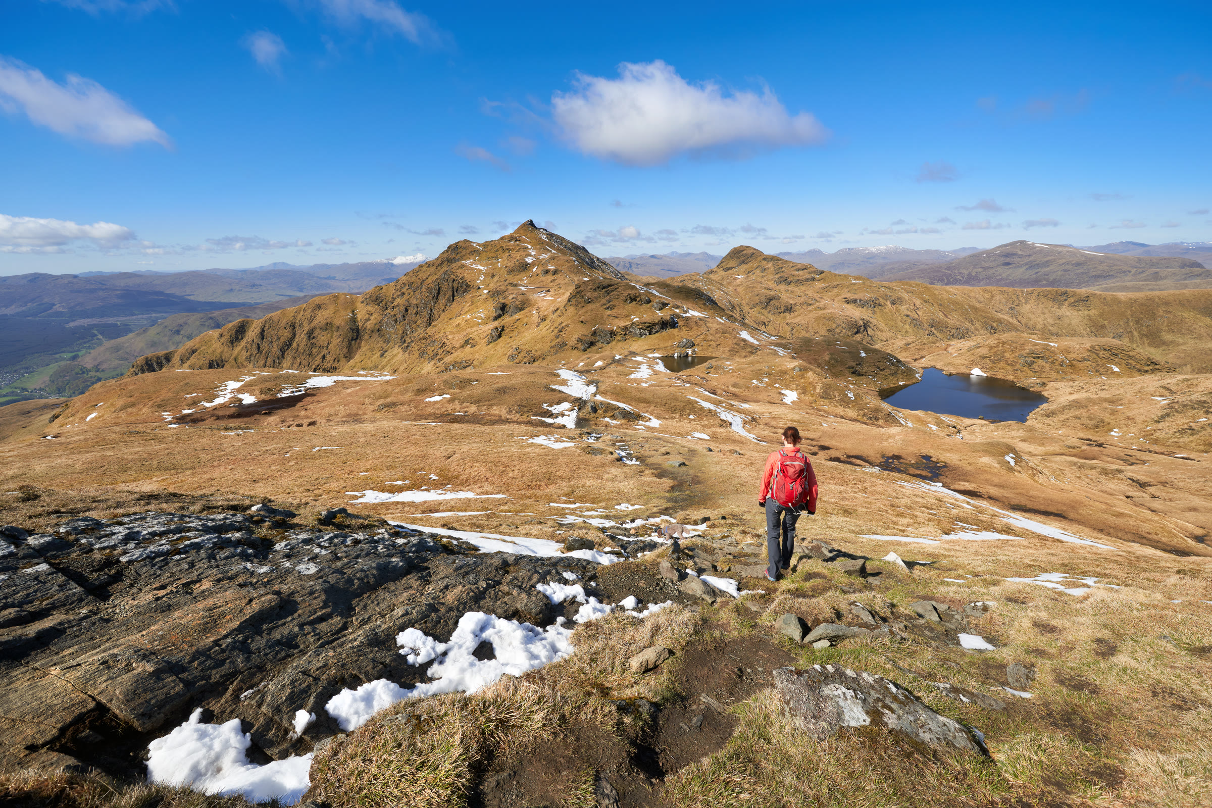 A walker looking along the Tarmachan ridge towards Meall Garbh and Beinn nan Eachan, a lochan below and patches of snow.
