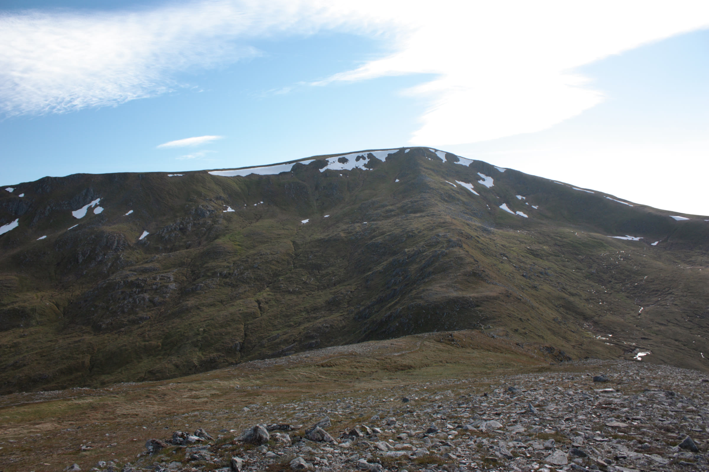 The broad top of Tom a' Chòinich with late snow patches, seen across stony ground.