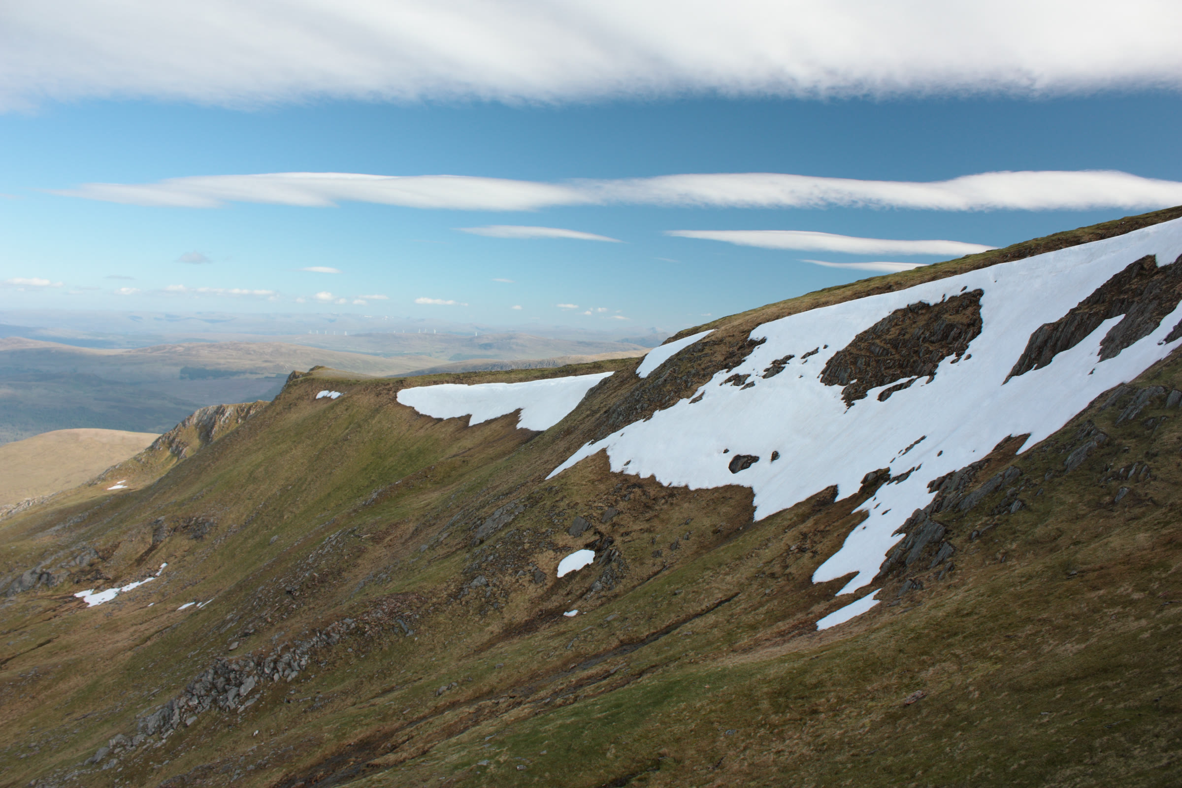 A large snowfield on the steep grassy slope of Tom a' Chòinich, under long bands of cloud.