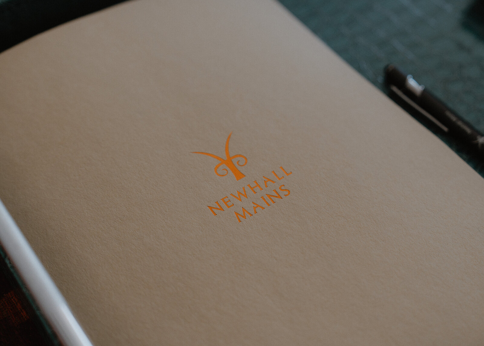 A cream card embossed with an orange goat's-head logo and the words Newhall Mains lies on a leather tray beside a pen.