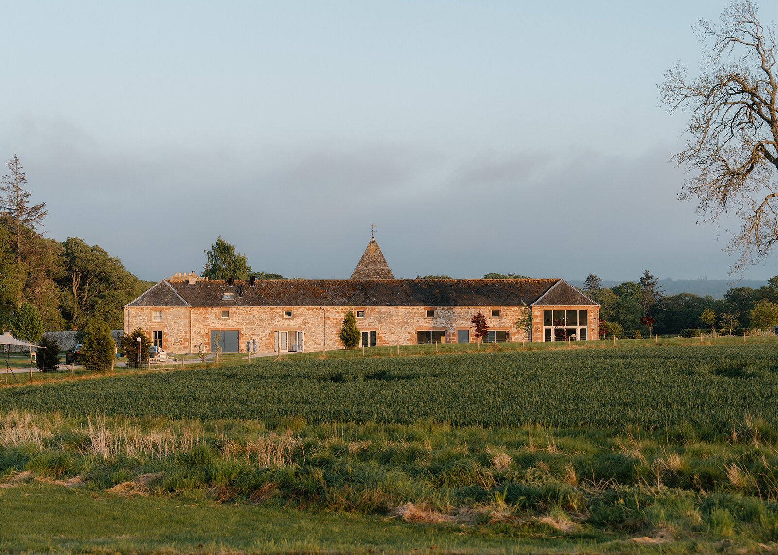 A long sandstone steading with a conical-roofed tower at its centre stands beyond a field of young crops, trees framing it on either side in evening light.