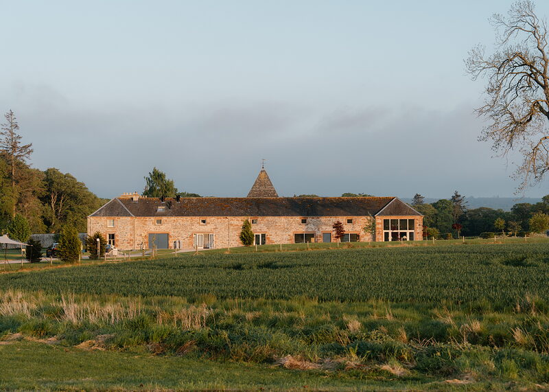 A long sandstone steading with a conical-roofed tower at its centre stands beyond a field of young crops, trees framing it on either side in evening light.
