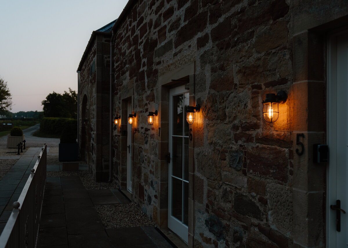 A row of wall lanterns lights a sandstone building at dusk, glazed doors numbered "5" standing along the path.