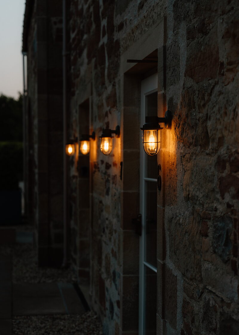 A row of lit wall lanterns lines a stone building at dusk, a white-framed doorway standing between them.