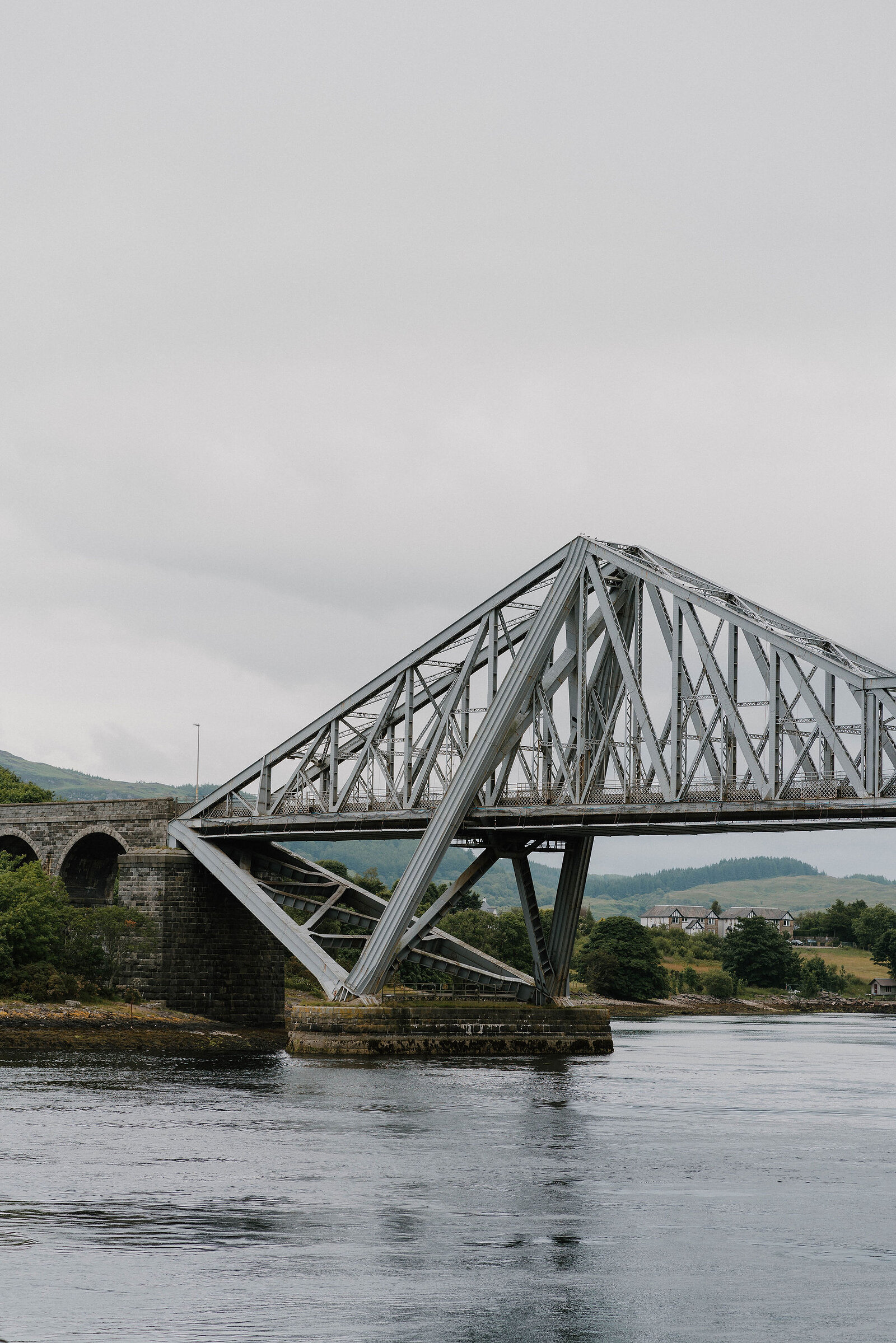 A steel lattice cantilever bridge spans a sea loch, hills and a scatter of houses visible beyond.