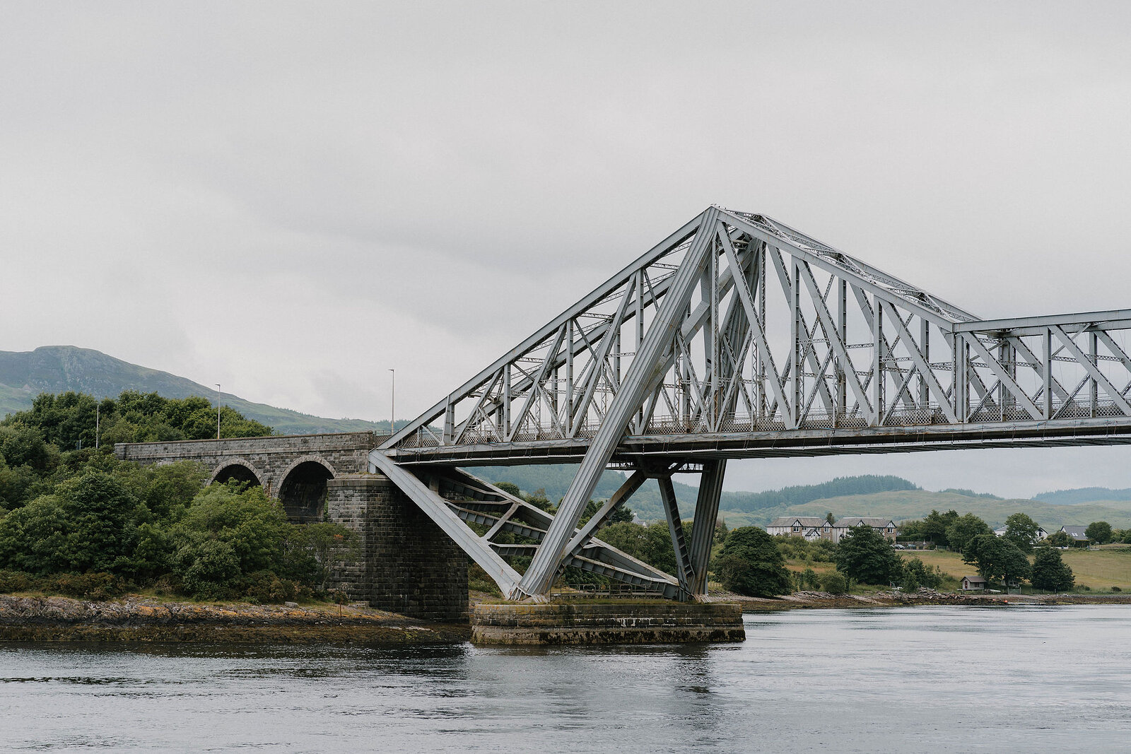 A steel cantilever bridge spans a river beside older stone arches, hills rising along the far bank.