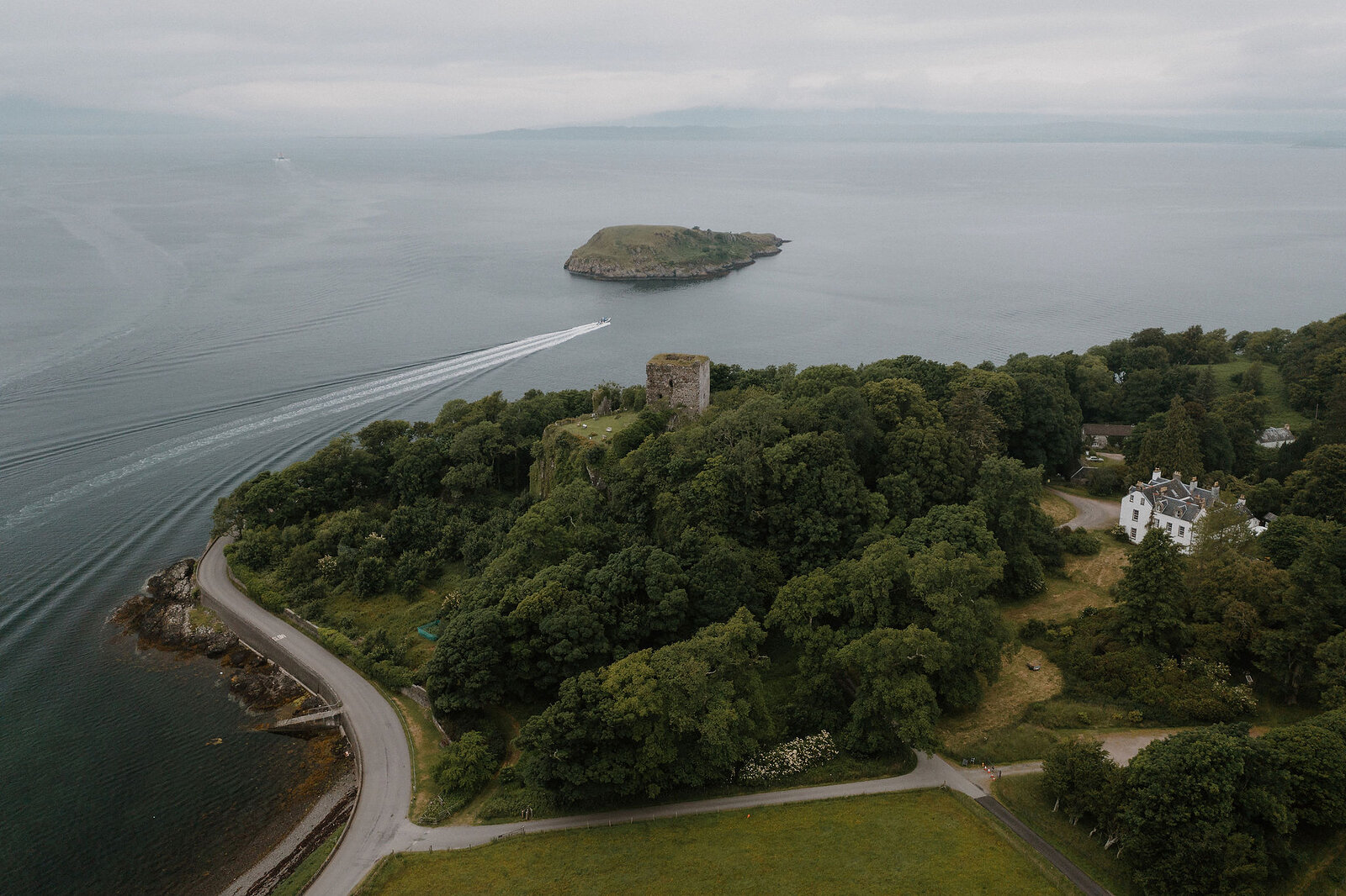 An aerial view shows a ruined tower rising from a wooded point beside a loch, a boat's wake curving past and a white mansion visible among trees beyond.