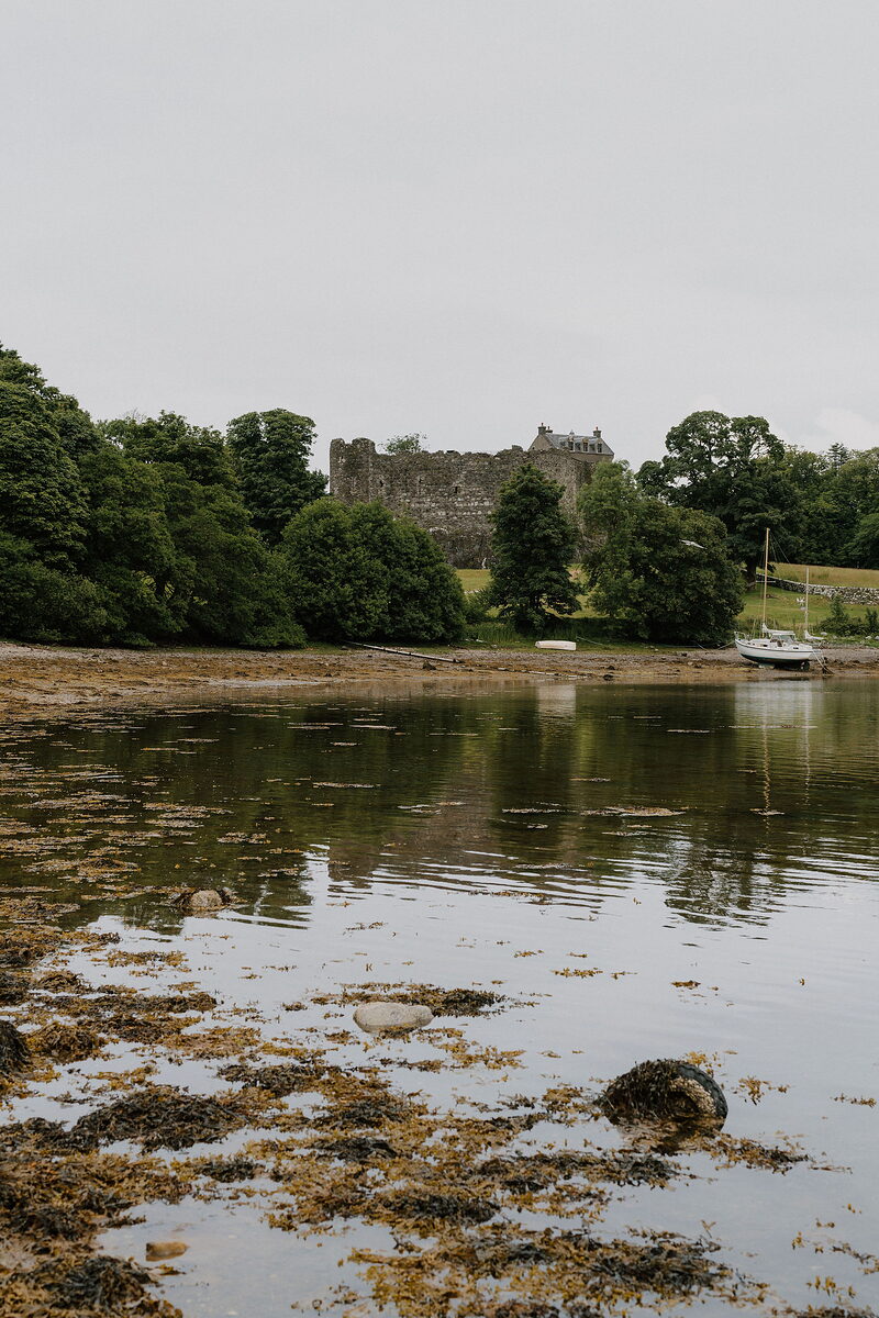 A ruined castle tower rises among trees on a wooded shore, a small boat moored nearby and its reflection mirrored in the still water.