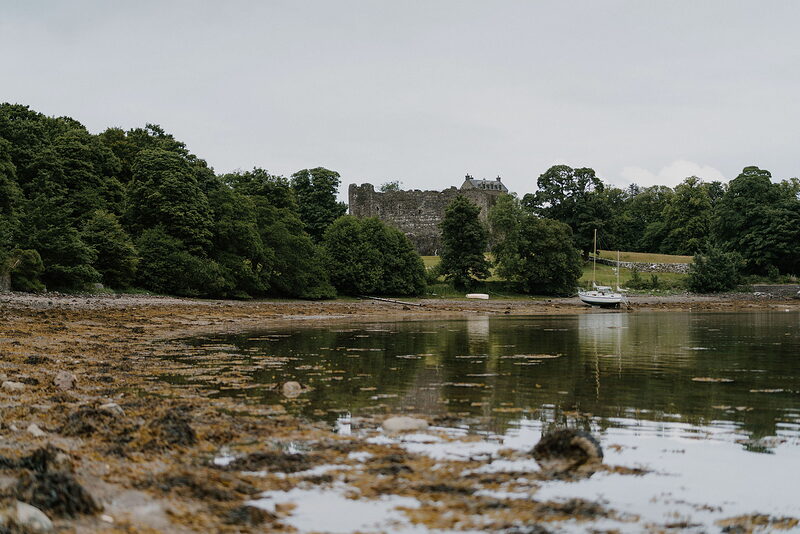A ruined castle tower rises among trees on a wooded shore, small boats moored nearby and their shapes mirrored in the still water.