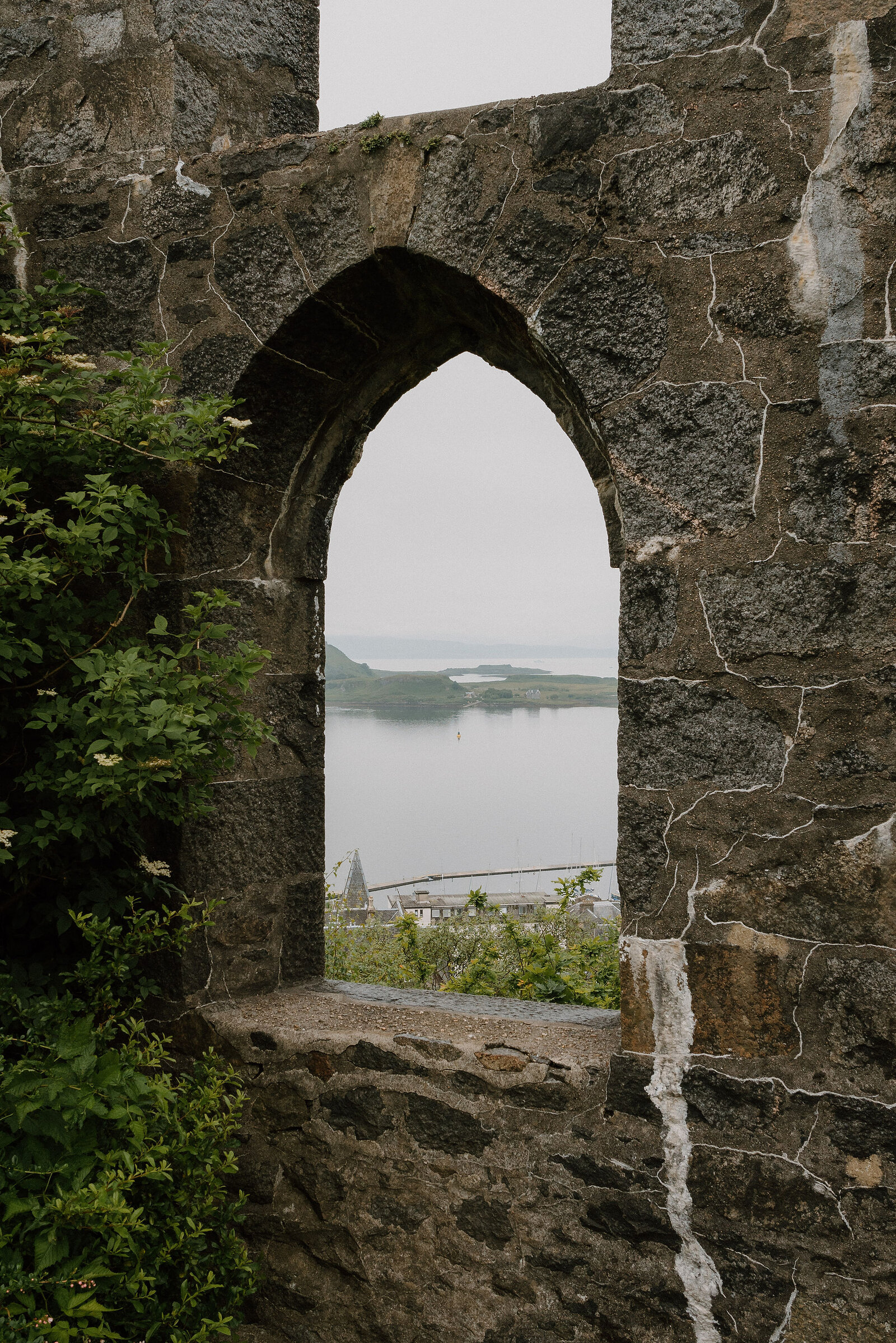 A pointed stone archway frames a view of Oban bay, a lighthouse and hills visible across the water with rooftops below.