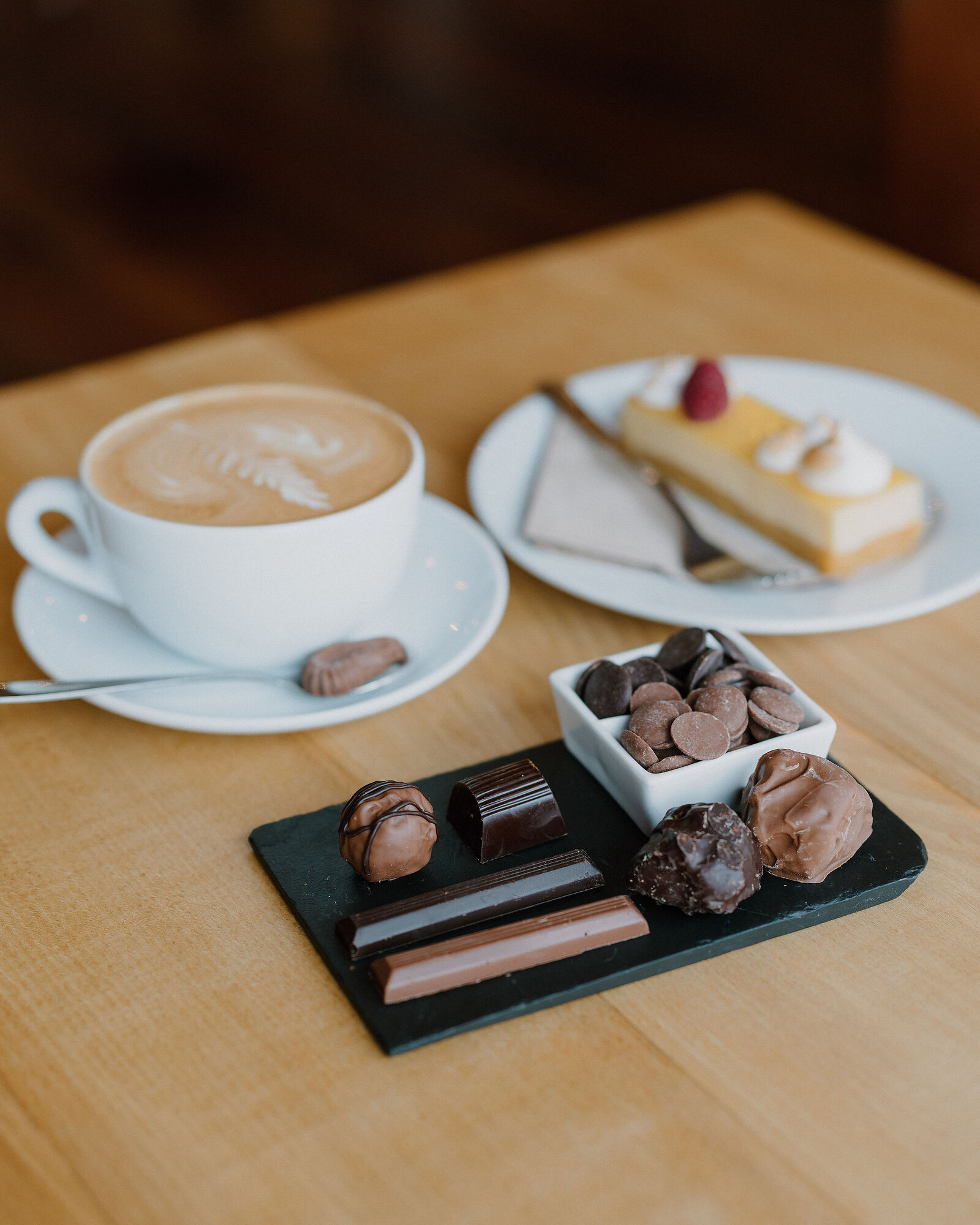 A latte and a slice of lemon tart topped with meringue and a raspberry sit on plates beside a small selection of chocolates on a slate board.