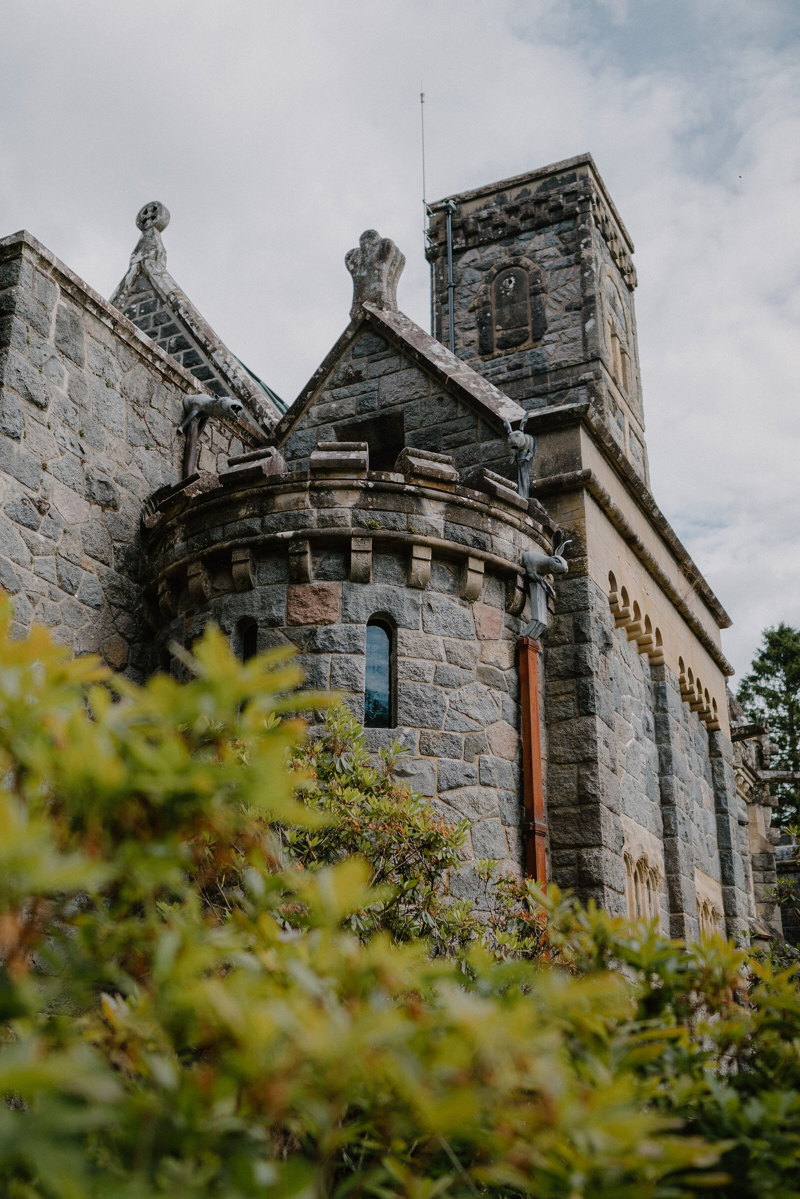 A stone Gothic chapel with carved gargoyles, a corner turret and a square bell tower rises behind green shrubs.