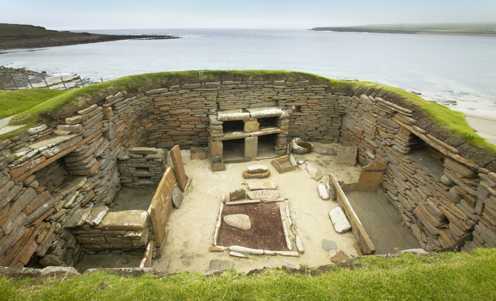 A stone house at Skara Brae seen from above, with its hearth and stone furniture, the Bay of Skaill behind.