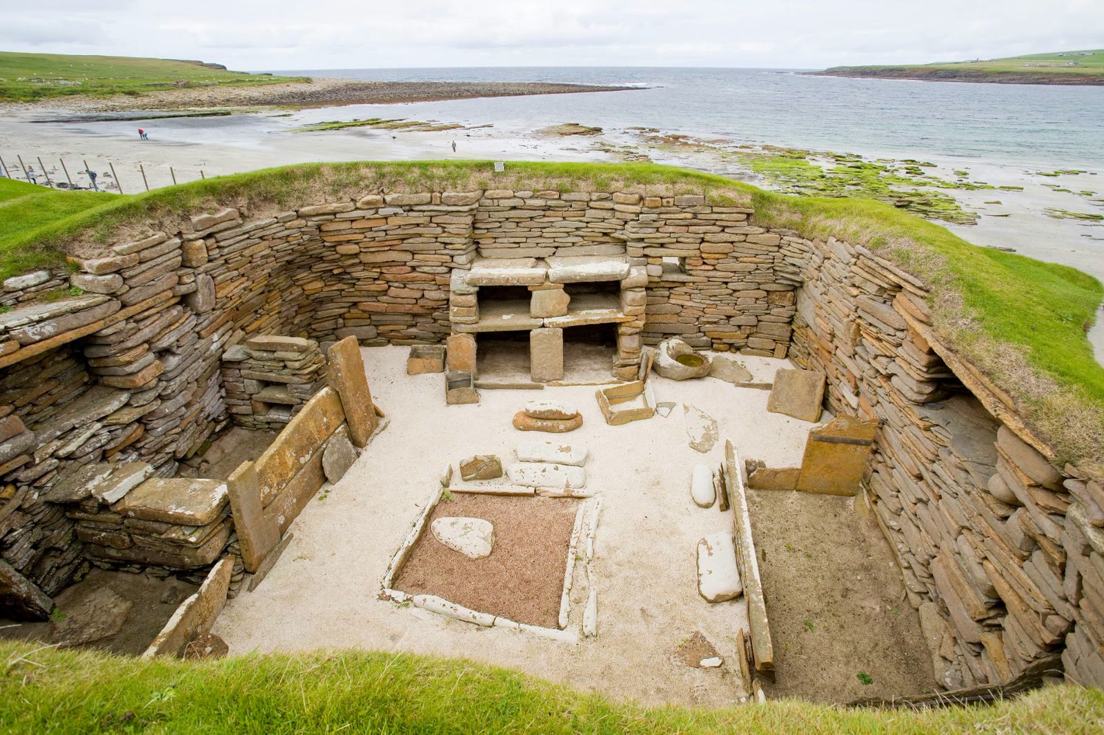 A house at Skara Brae with the sand of the Bay of Skaill behind it and people walking on the beach.