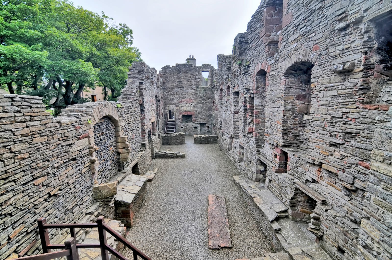 Inside the roofless palaces in Kirkwall, a long range of flagstone walls with arched recesses either side of a gravel floor running to a stair, Orkney.