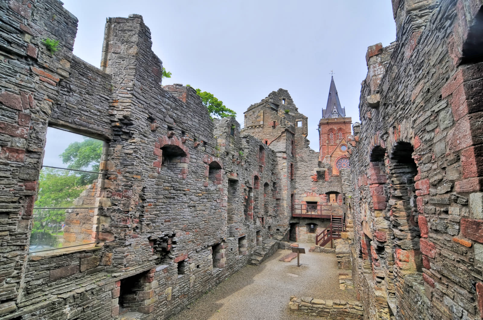 Inside the roofless palaces in Kirkwall, grey and red stone walls with deep window openings, the spire of St Magnus Cathedral rising beyond, Orkney.