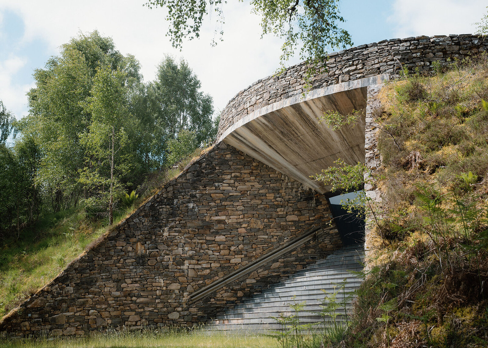 A wider side view of the skyspace entrance, its drystone wall and slate steps set into a heather bank with birches behind.