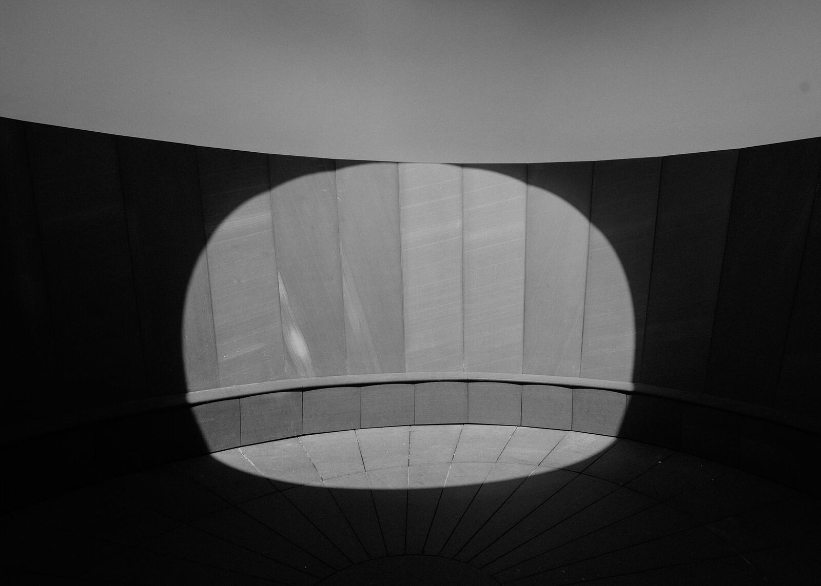 In black and white, a rounded patch of sunlight falls on the chamber's panelled wall, bench and radiating floor, the rest in shadow.