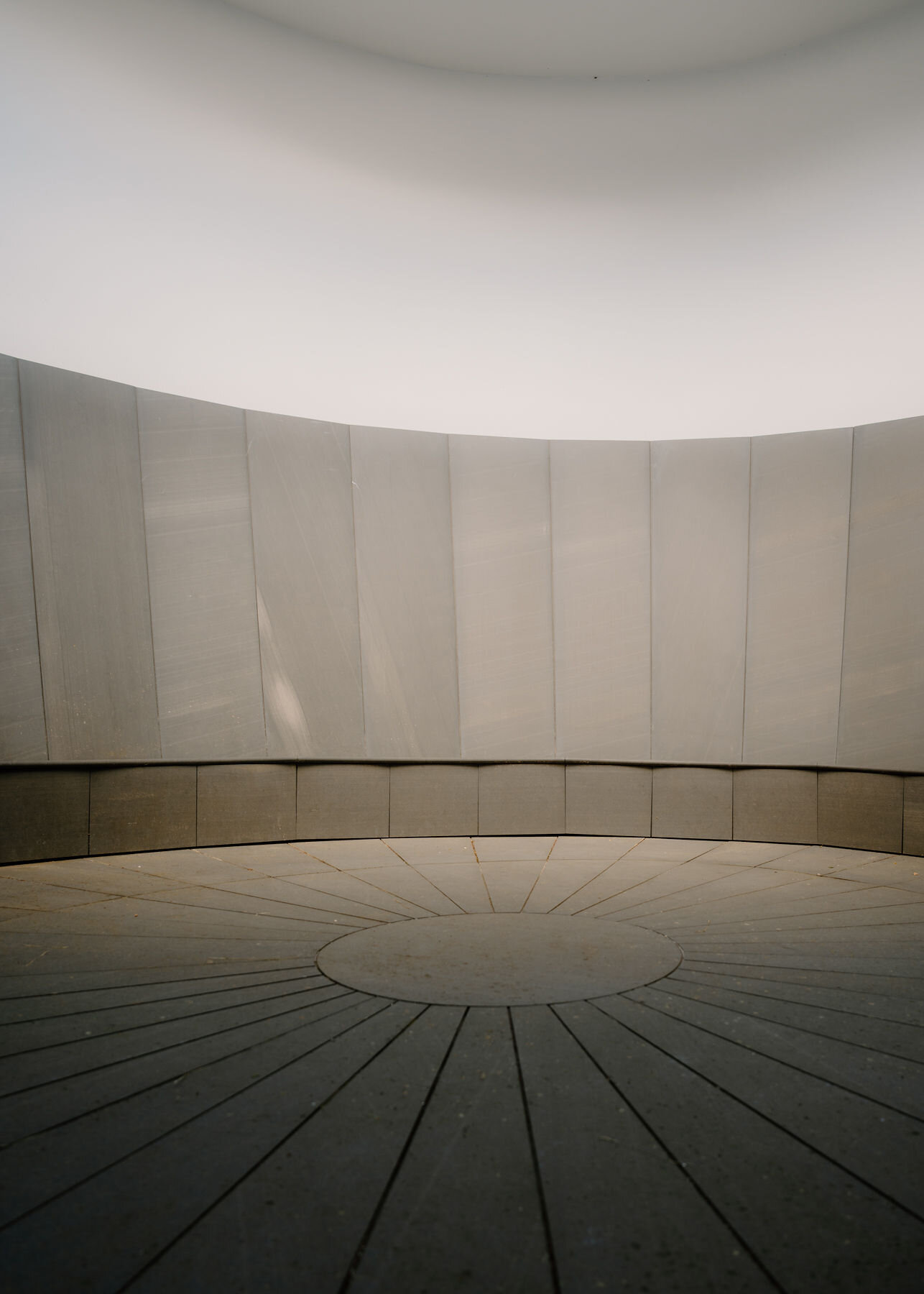 Inside the skyspace chamber, a stone bench runs round the panelled wall above a floor of radiating slabs with a central disc.