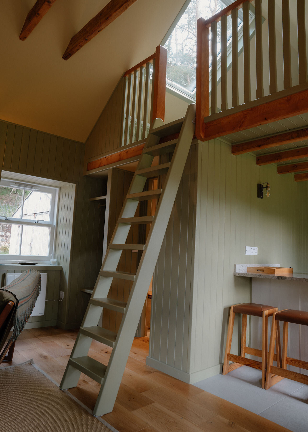 A green-painted ladder climbs to a sleeping loft with a timber balustrade, lit by a roof window, beside a sash window and two bar stools.