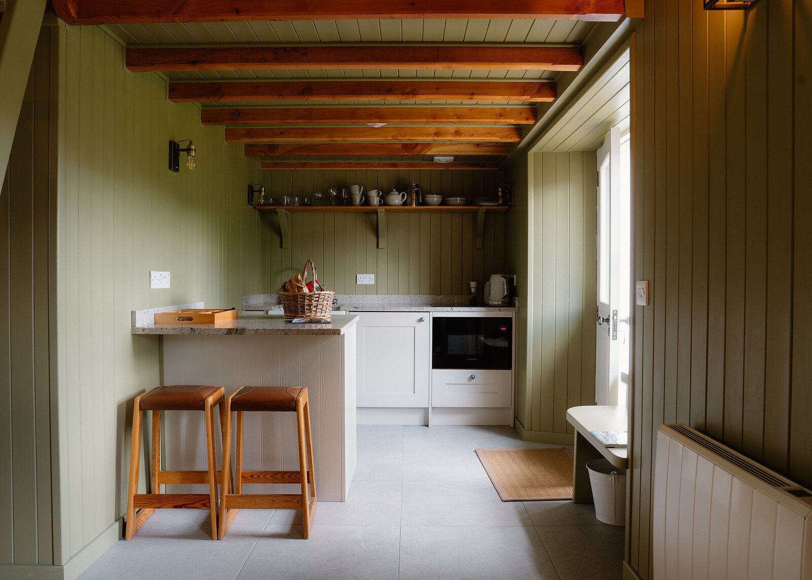 Timber beams run the length of a green-panelled kitchen, with two leather-topped stools at the bar, a bench by the door and a radiator.
