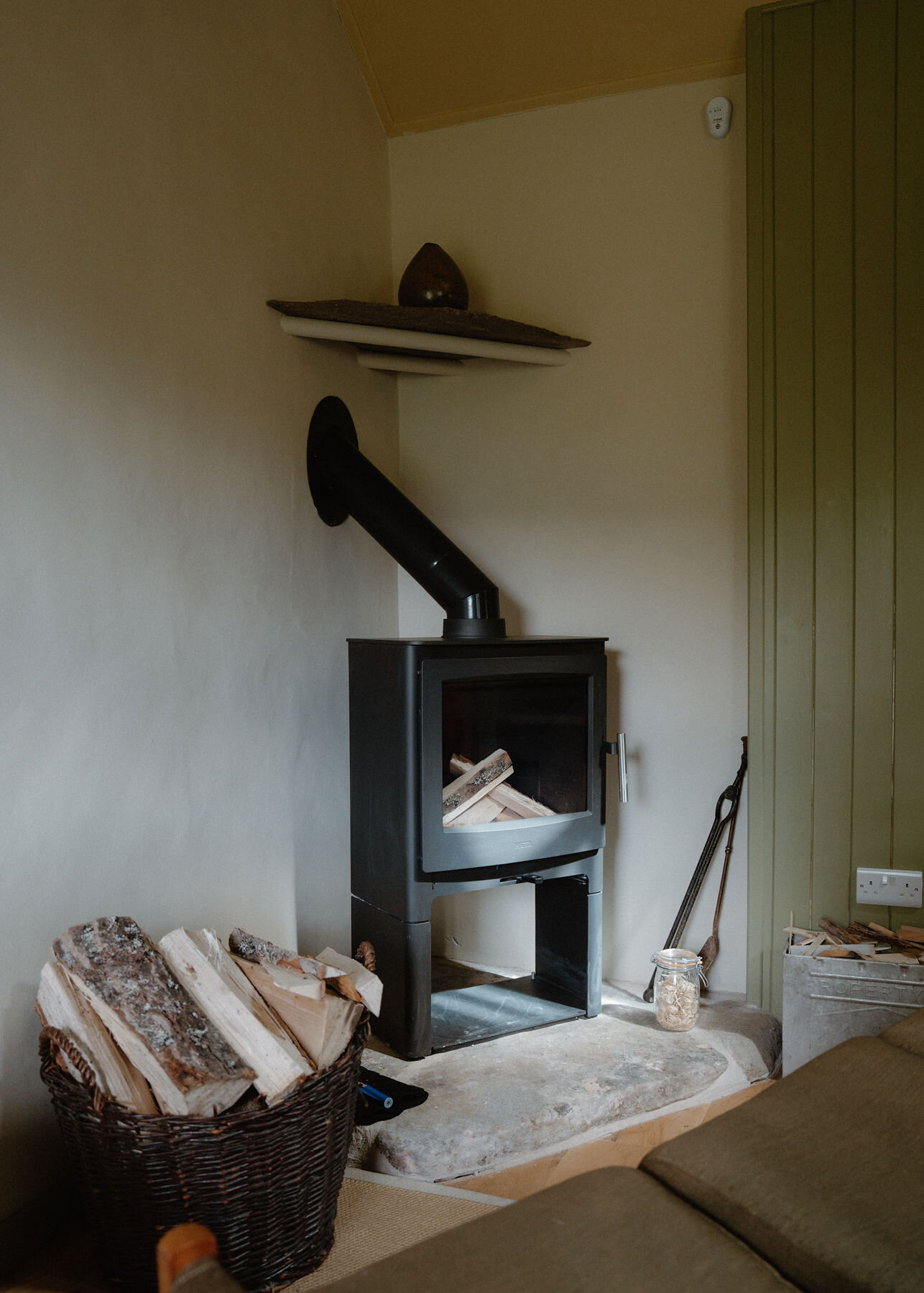 A black wood-burning stove, unlit and laid with logs, stands on a stone hearth beside a wicker log basket, with a slate shelf above.