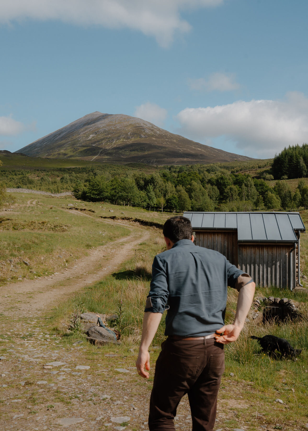 A man walks towards a timber hut below Schiehallion's cone, a black dog nosing the grass and a child hiding among the rocks.