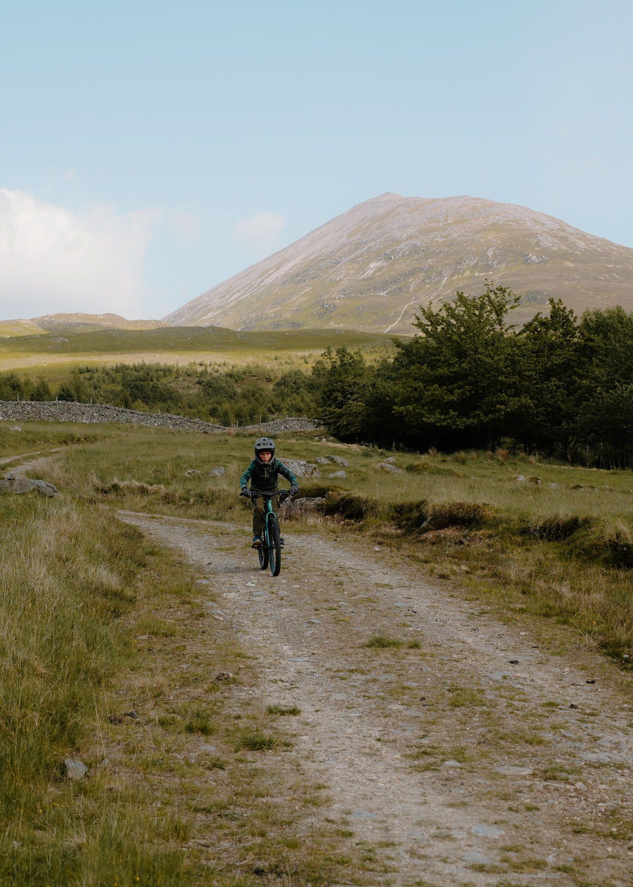 A boy in a full-face helmet rides a mountain bike down a stony farm track, with Schiehallion's cone rising beyond a belt of trees.