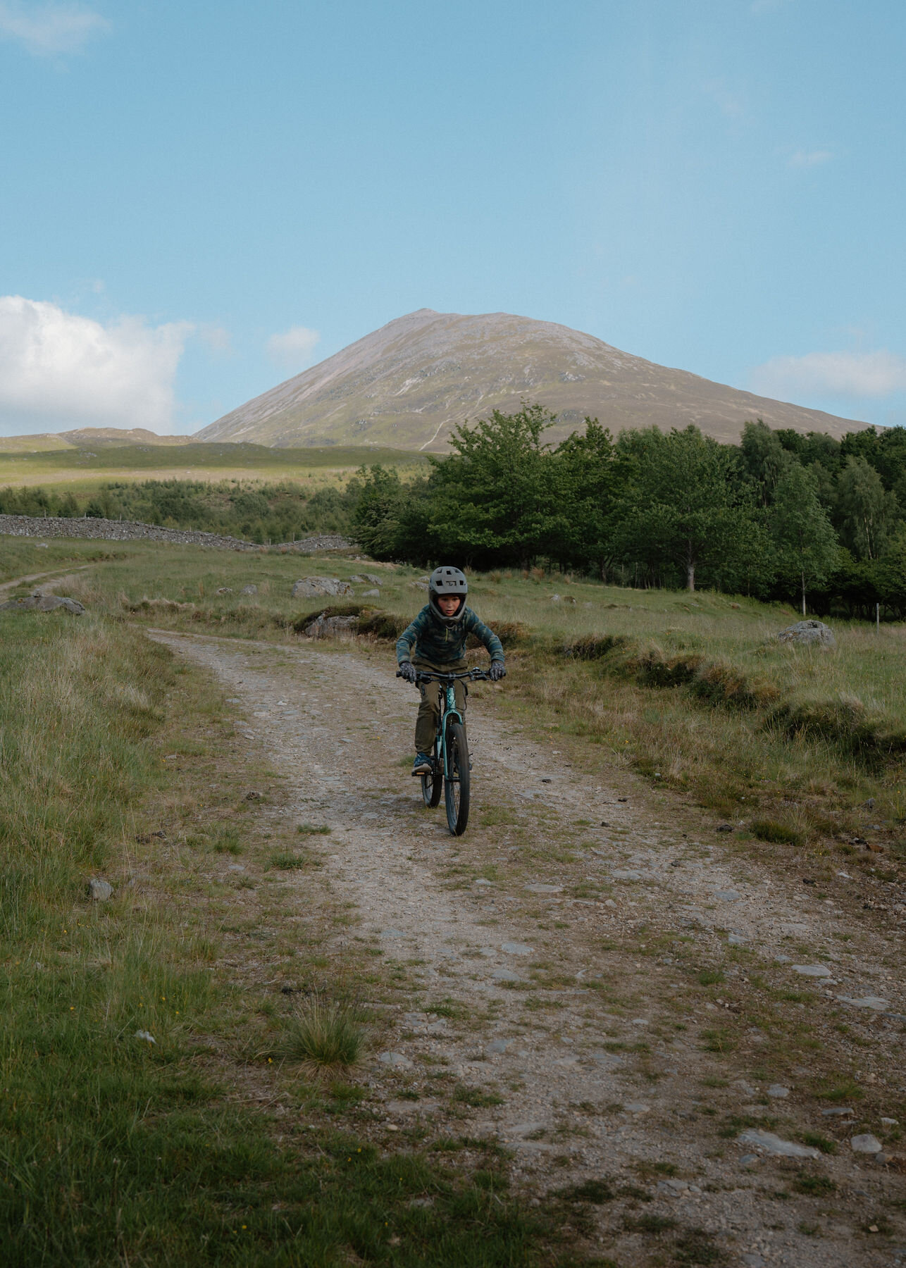 A helmeted boy on a mountain bike comes closer down the gravel farm track, Schiehallion standing under a wide blue sky behind.