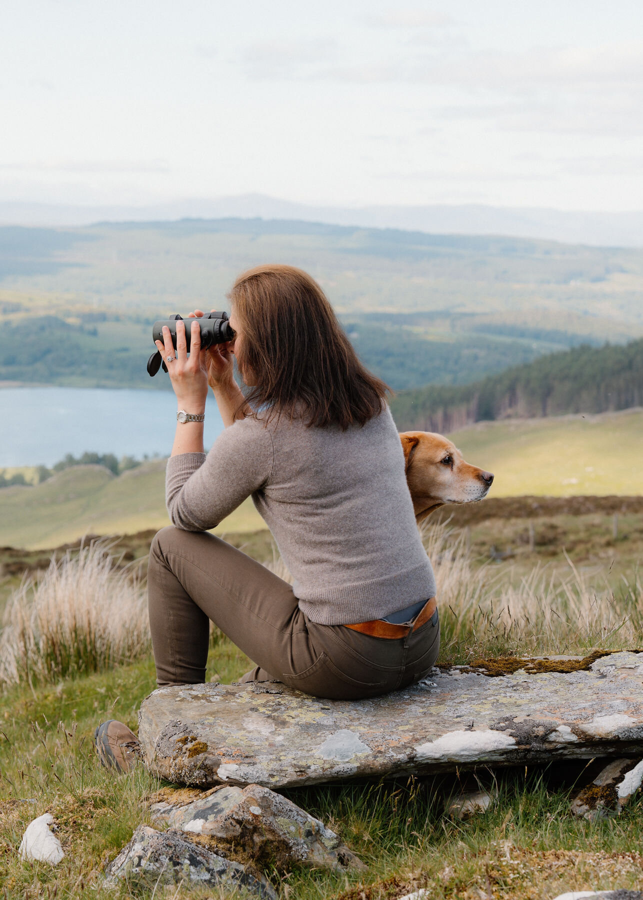 A slightly wider view of a woman on a lichened rock raising binoculars towards the loch, a golden dog behind her gazing the other way.