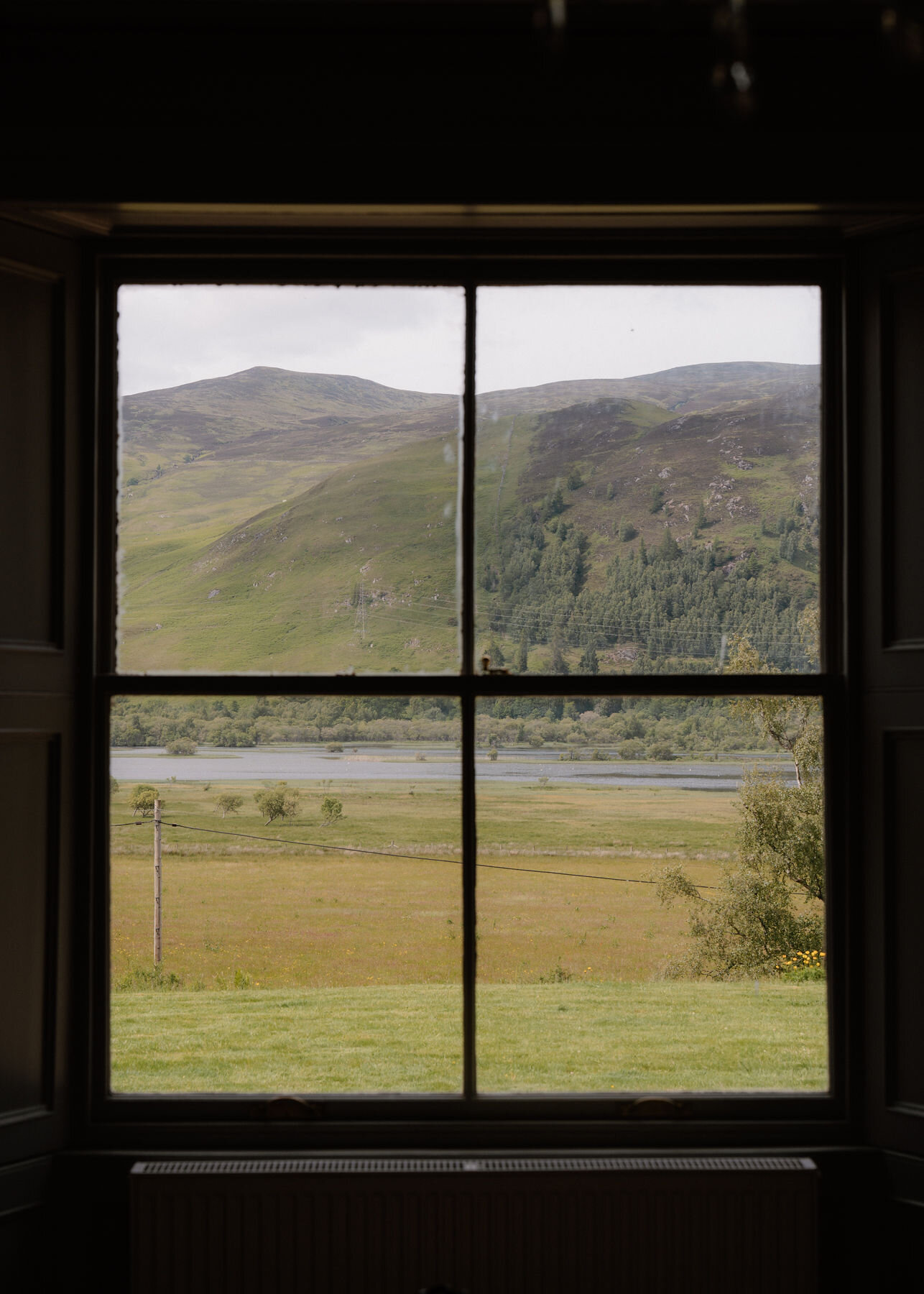 A dark sash window frames a green hillside above a loch and river flats, with rough grass and a fence in front.