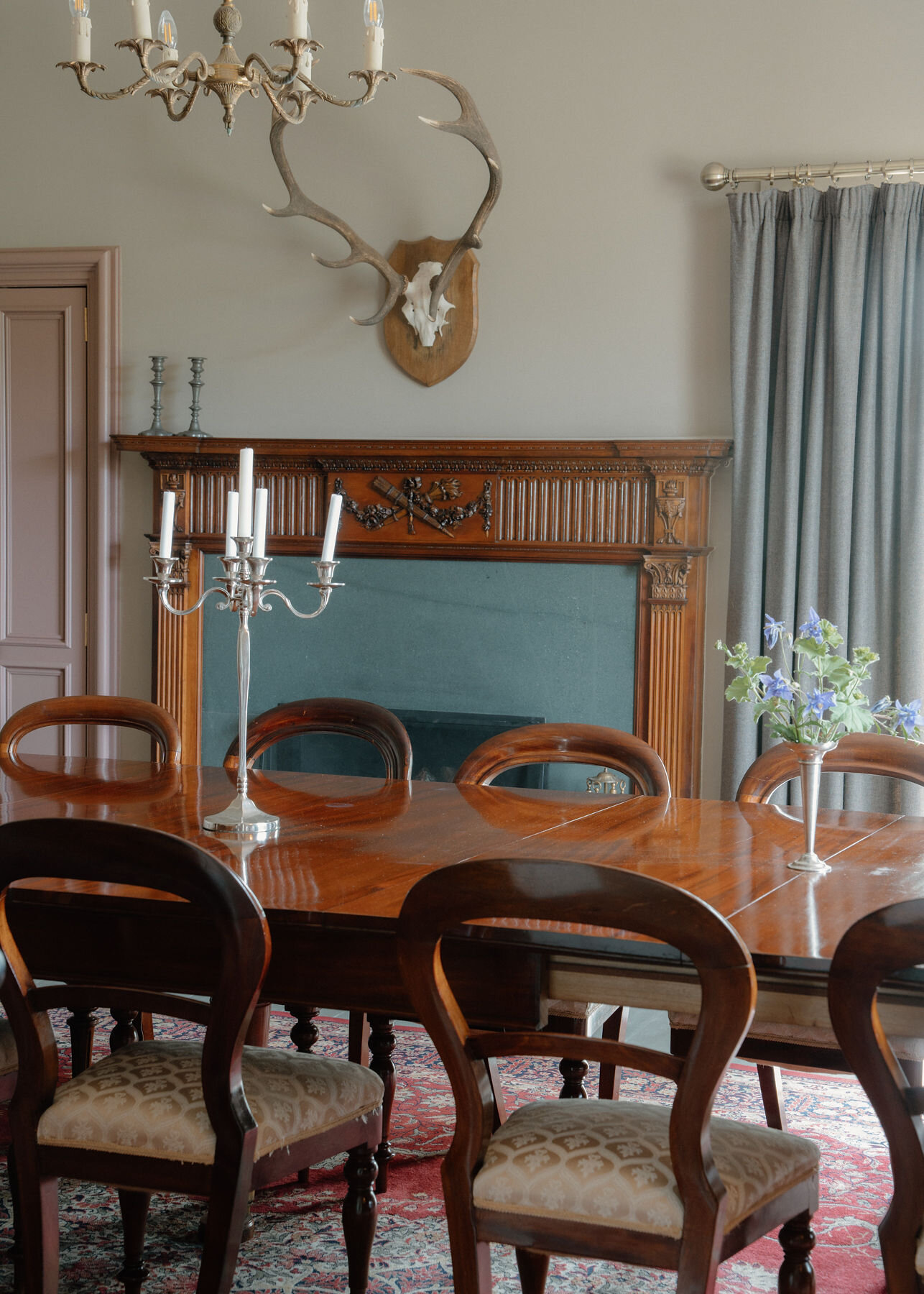 A mahogany dining table with balloon-back chairs stands under a candelabra, a mounted stag's antlers above a carved fireplace.