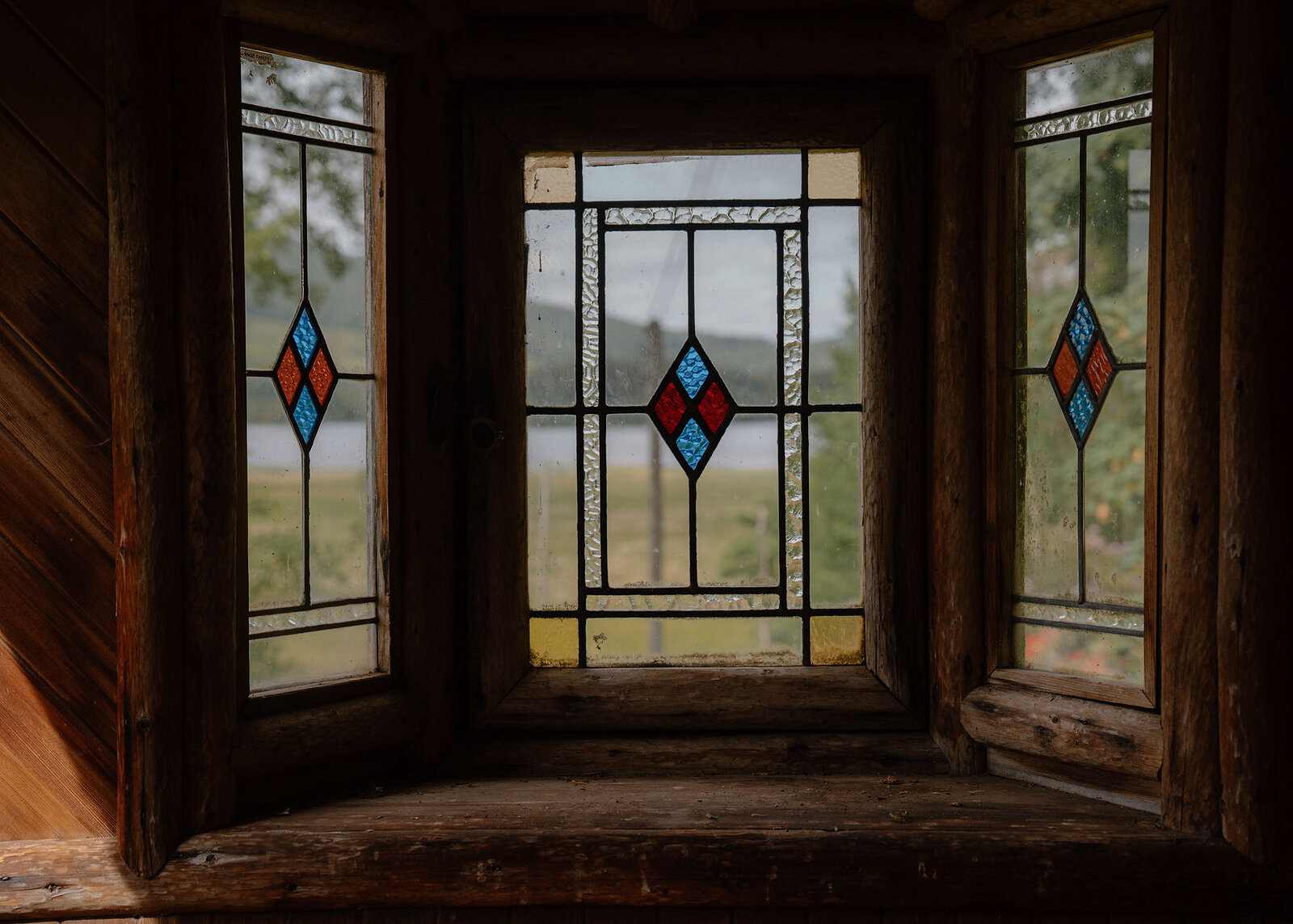 Three stained-glass windows with red and blue diamonds light a wood-panelled bay, the loch faint beyond the glass.