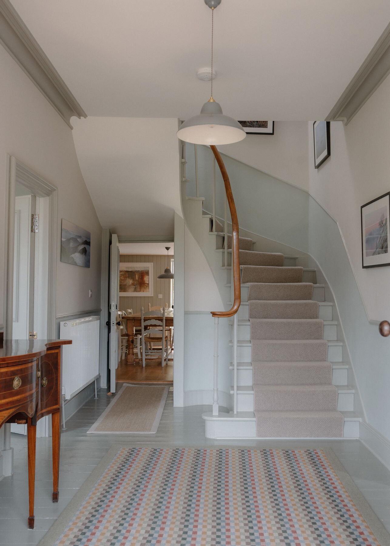 A bright hallway with a patterned runner leads to a curving staircase, a side table on the left and a dining room glimpsed beyond.