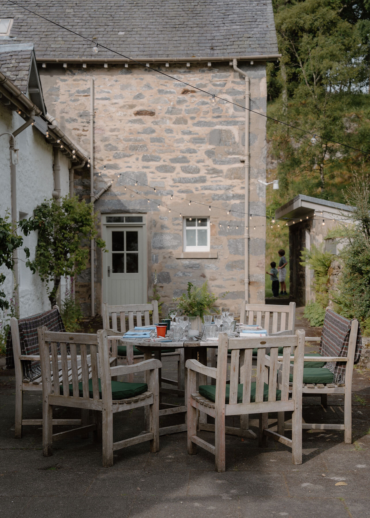 A round teak table laid for dinner with green-cushioned chairs and plaid throws on a patio under festoon lights, two children by a stone building.