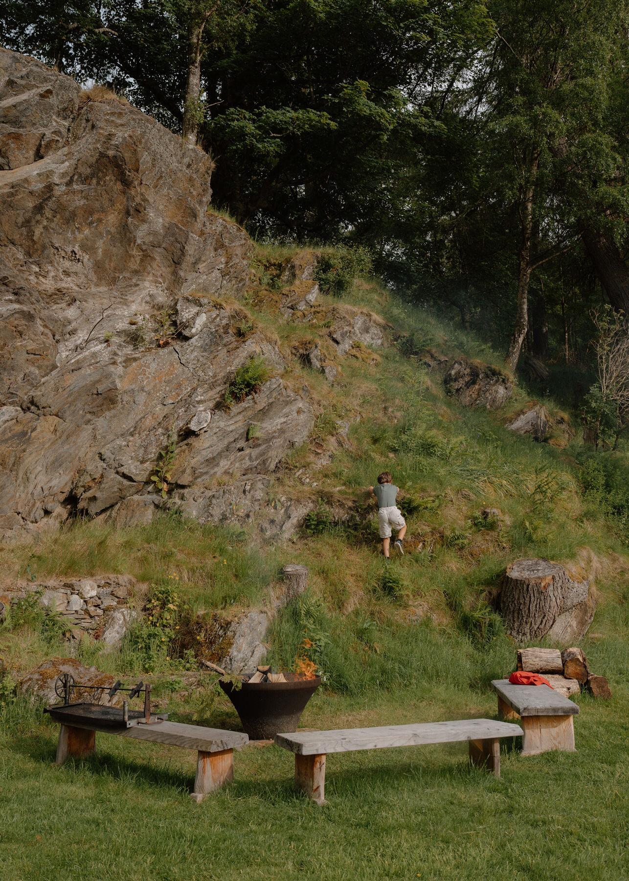 A boy climbs the grassy rock slope above a lit fire bowl, benches, a grill, a log pile and a tree stump.