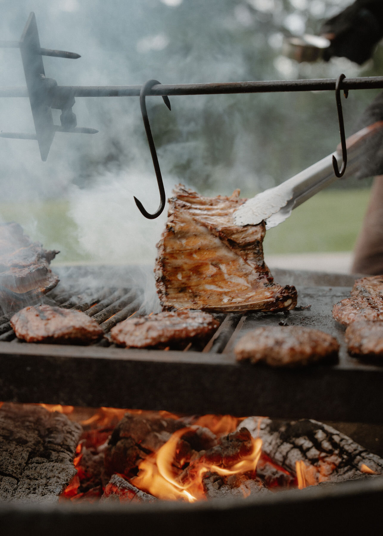 A rack of ribs held upright in tongs above the embers of the open-fire grill, burgers and hooks beside.